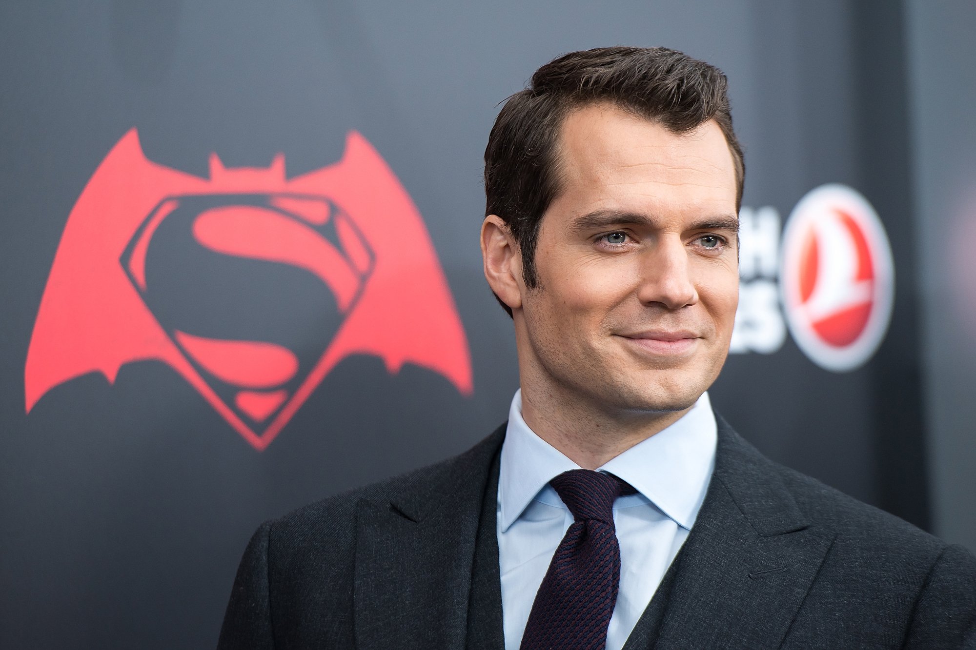Henry Cavill wearing a suit and standing in front of a wall with the Superman logo. Could Henry Cavill play Captain Britain in the Marvel Cinematic Universe?