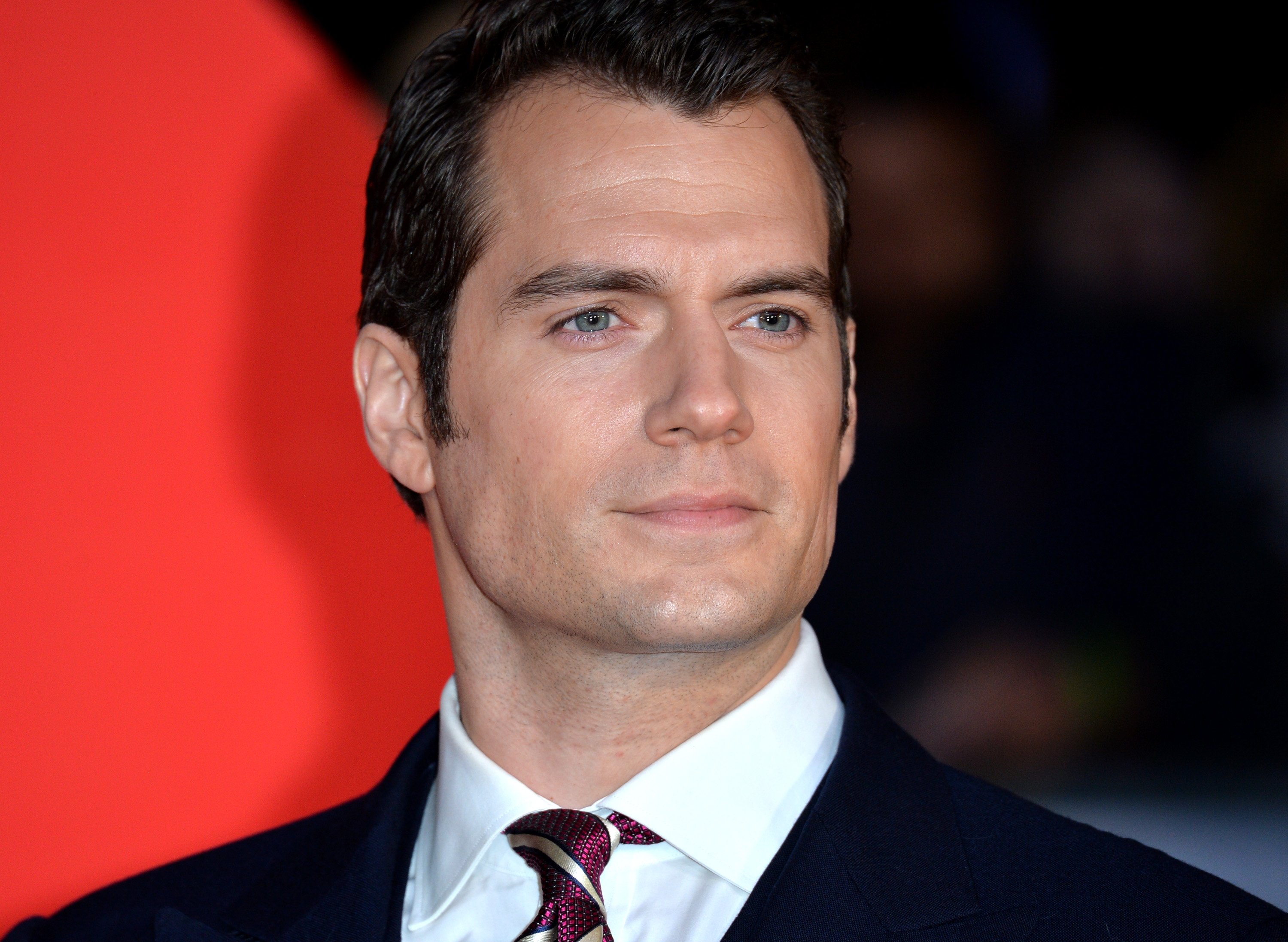 Henry Cavill, who plays Superman in the DC Extended Universe, wears a black suit over a white shirt and red tie.