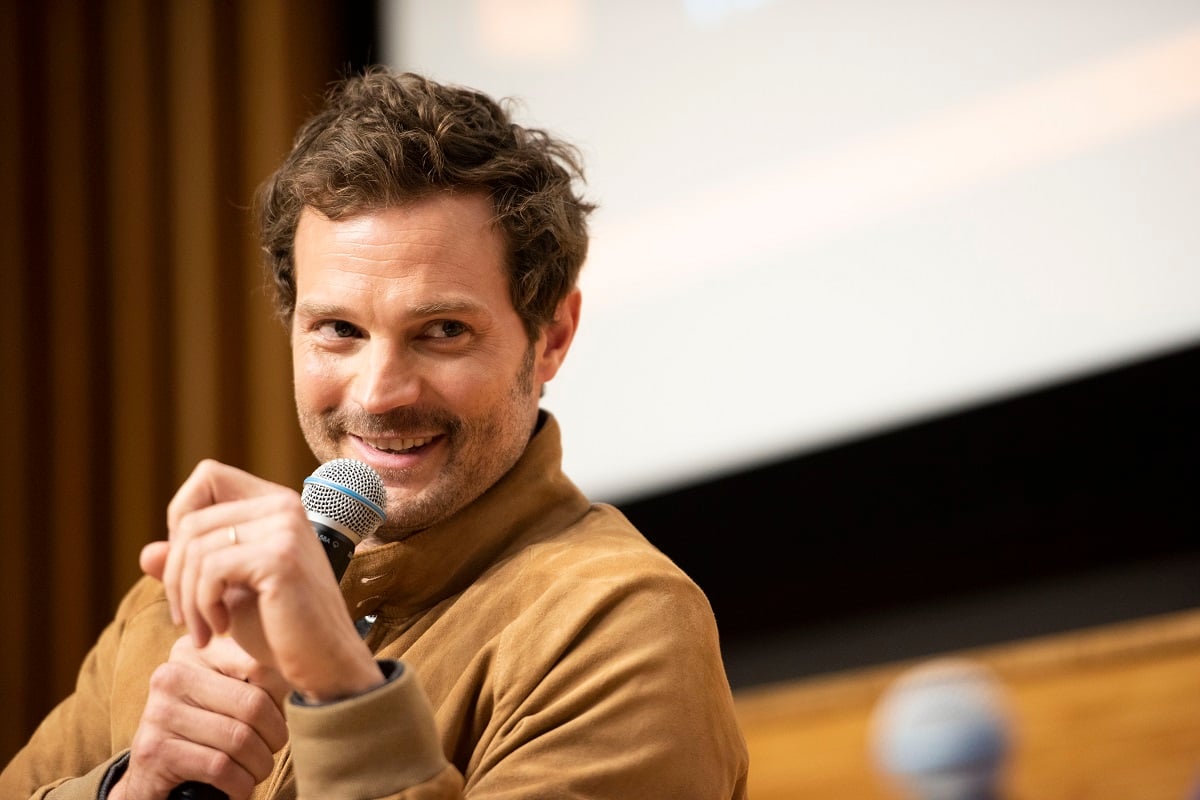 Jamie Dornan smiling in a brown sweater while holding a microphone