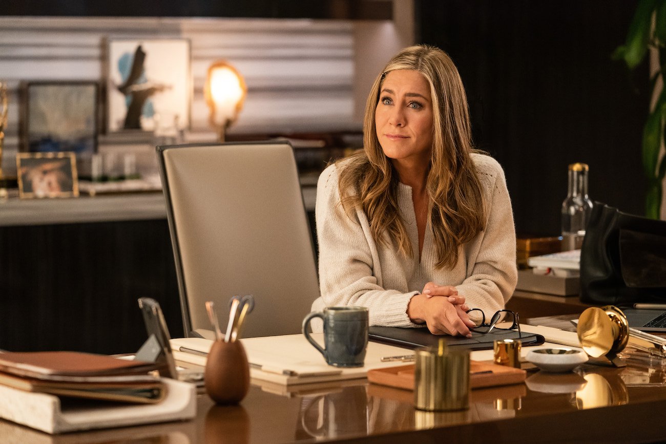 Jennifer Aniston sits with her hands on a desk and looks on