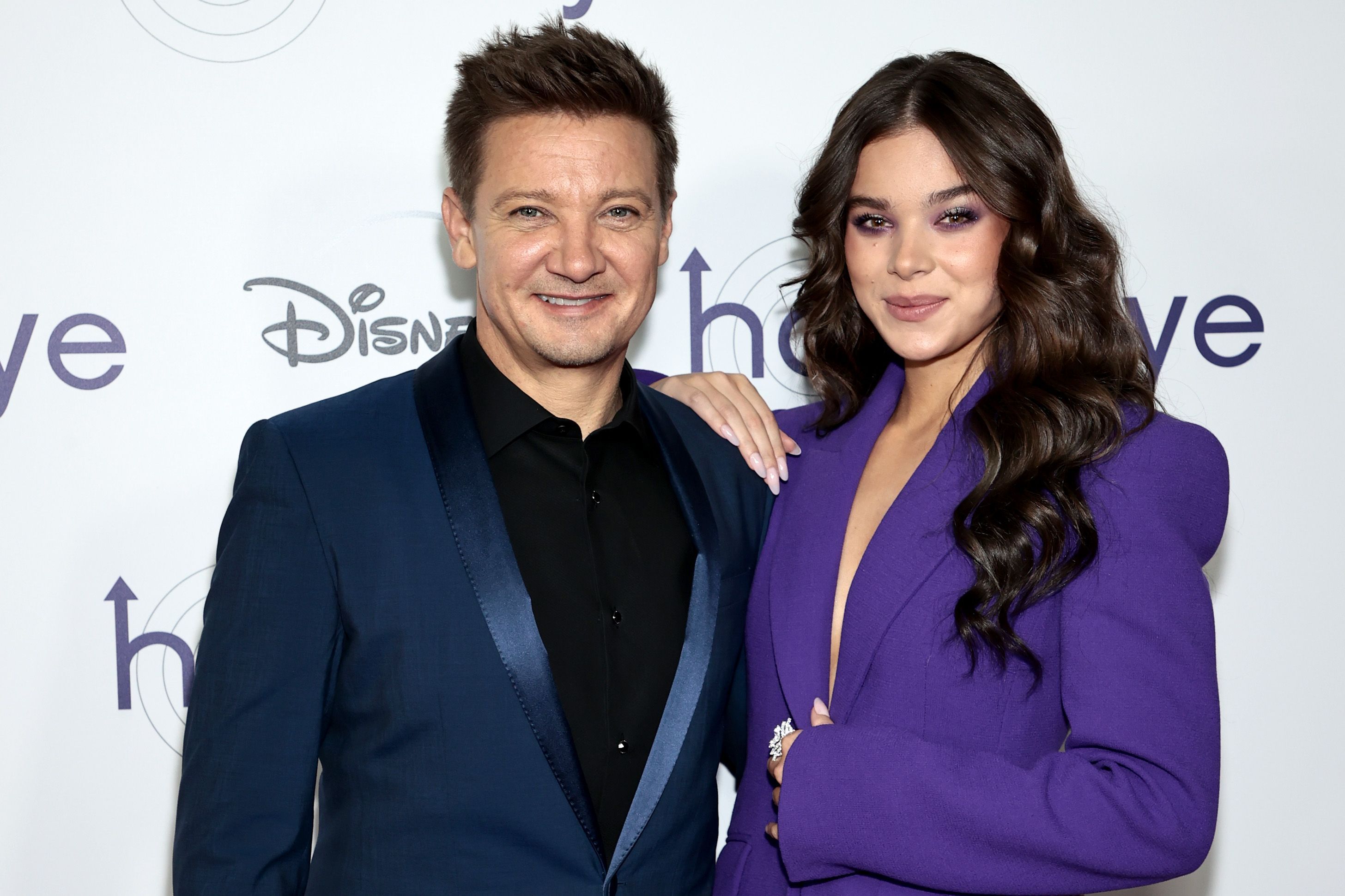 'Hawkeye' stars Jeremy Renner and Hailee Steinfeld pose for a picture together. Renner wears a dark blue suit over a black button-up shirt. Steinfeld wears a purple suit.