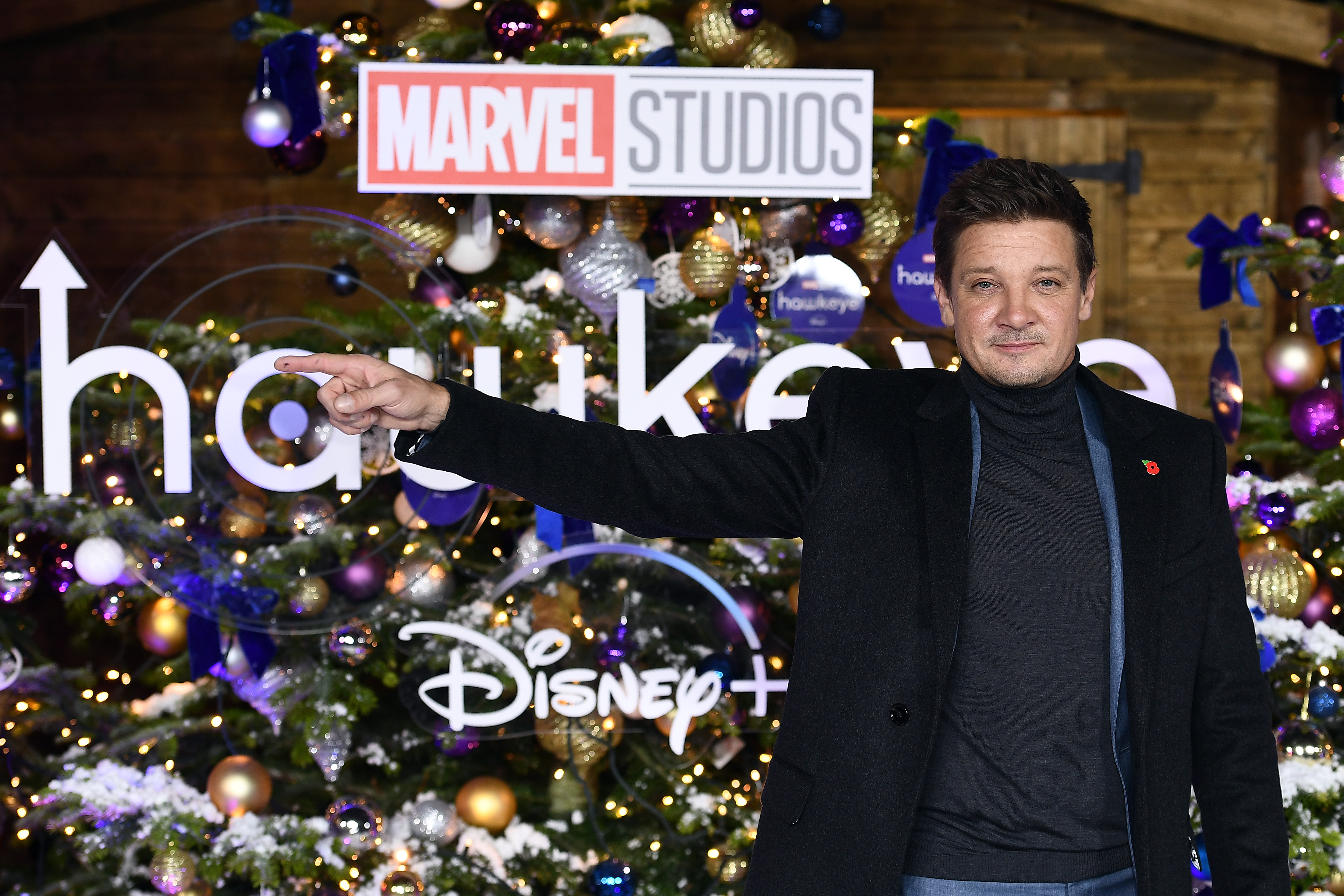 'Hawkeye' star Jeremy Renner, who plays Clint Barton, poses in front of a sign for his upcoming Marvel Disney+ show and a Christmas tree. Renner wears a black coat over a dark gray turtleneck sweater.