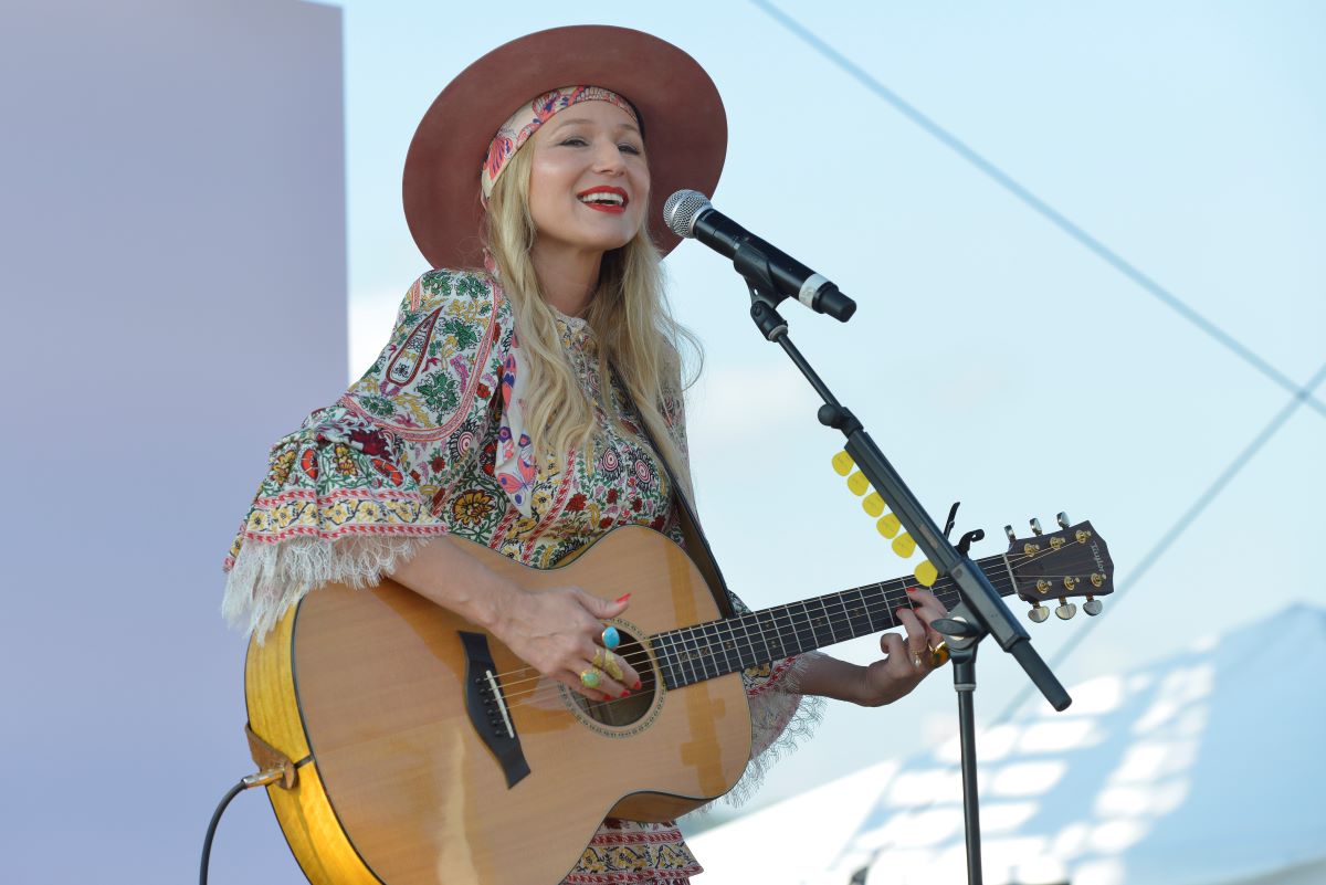 Jewel performs wearing a floral dress and hat holding a guitar and singing into a microphone