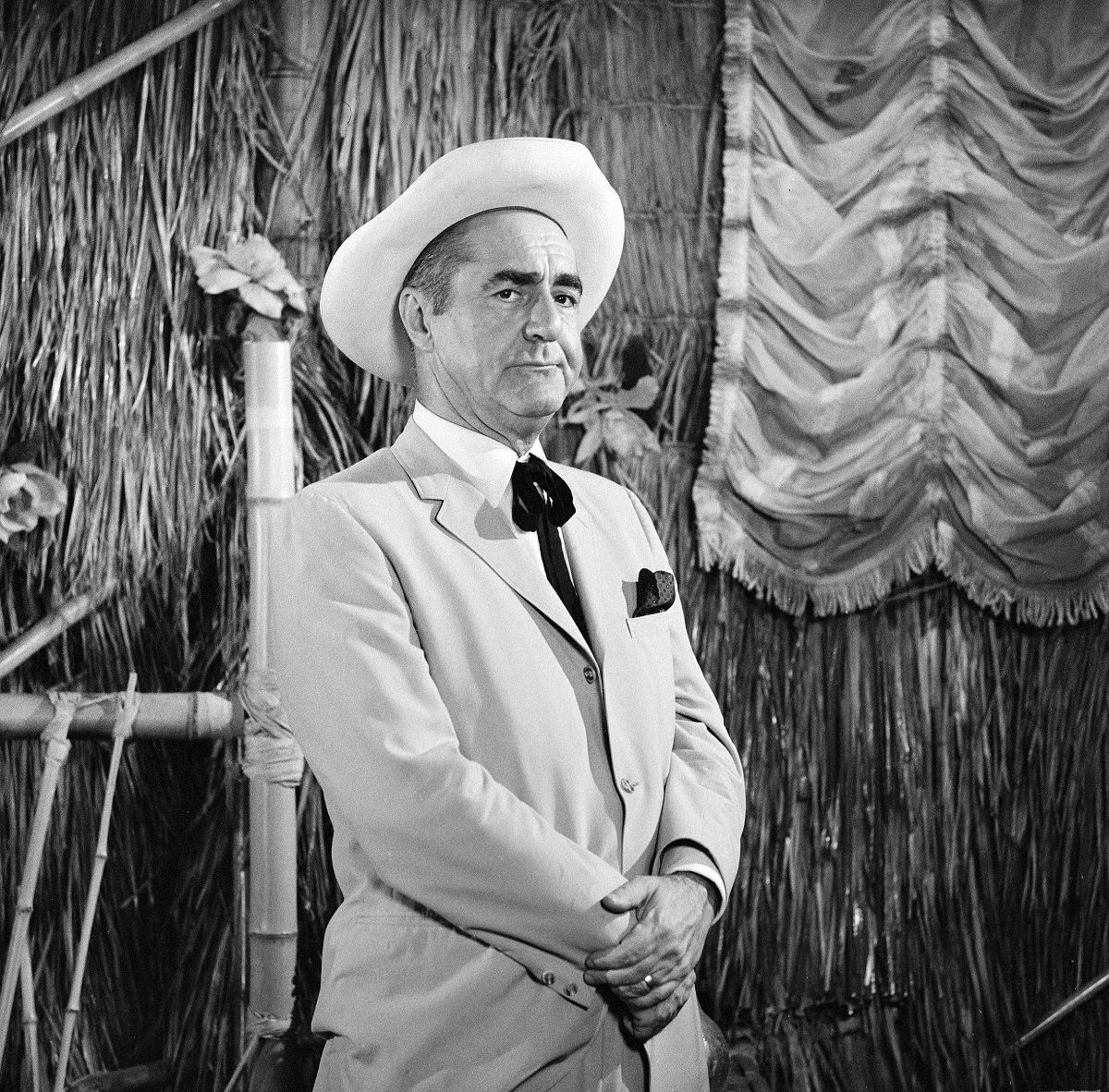 'Gilligan's Island' star Jim Backus wearing a suit and hat, standing in front of a bamboo hut.