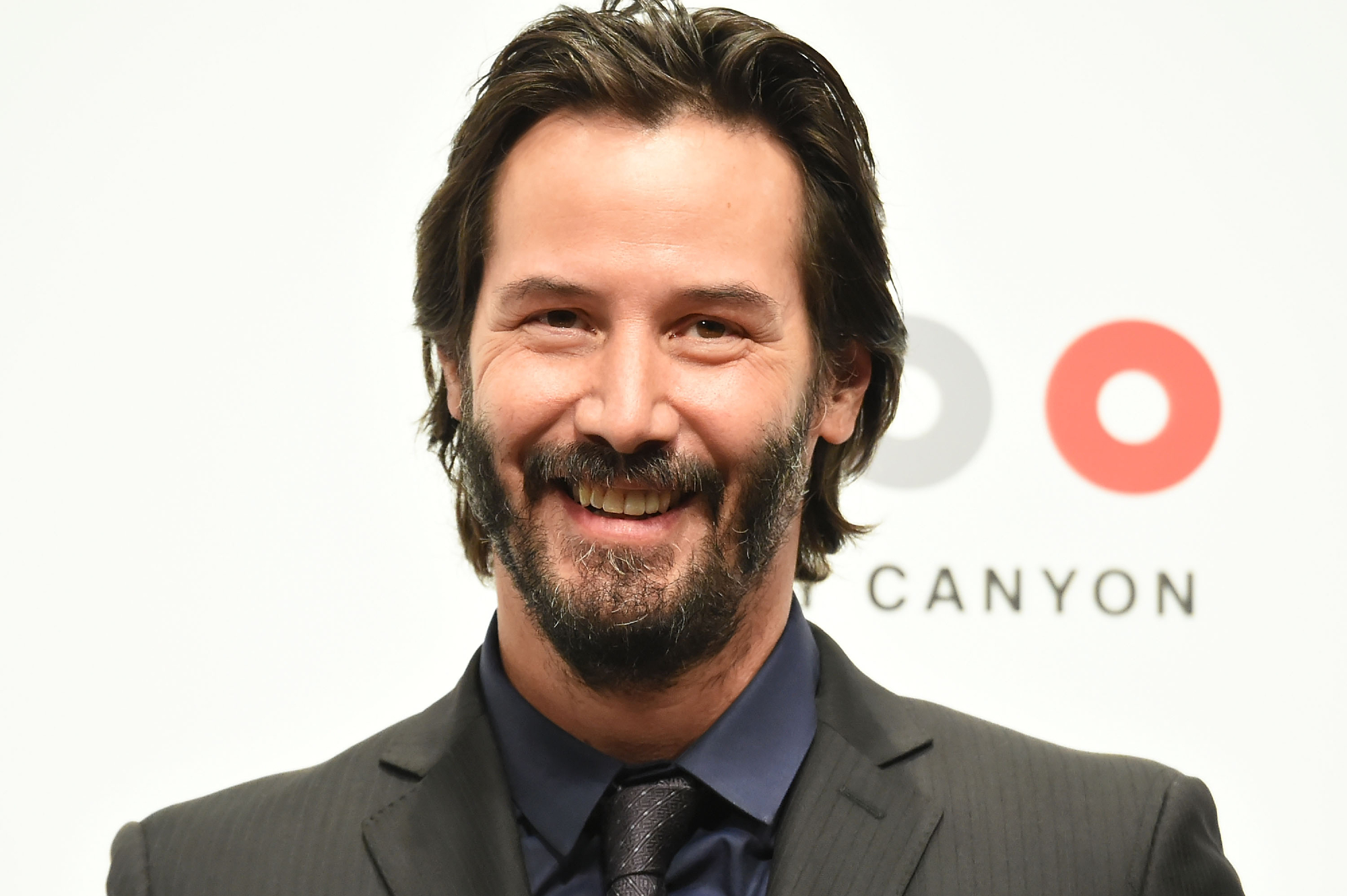 'John Wick 4' star Keanu Reeves smiling in a black suit in front of a step and repeat
