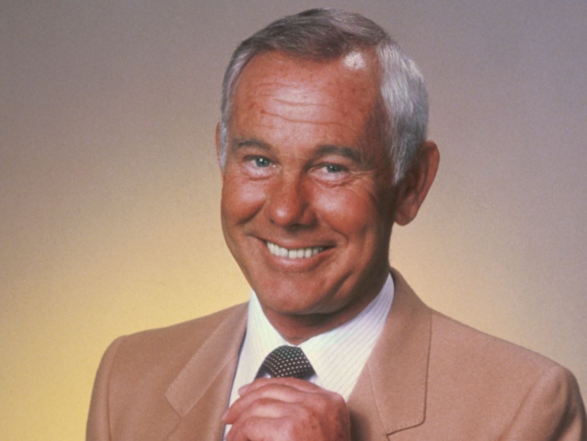 Johnny Carson smiles and holds his tie in a tan jacket  