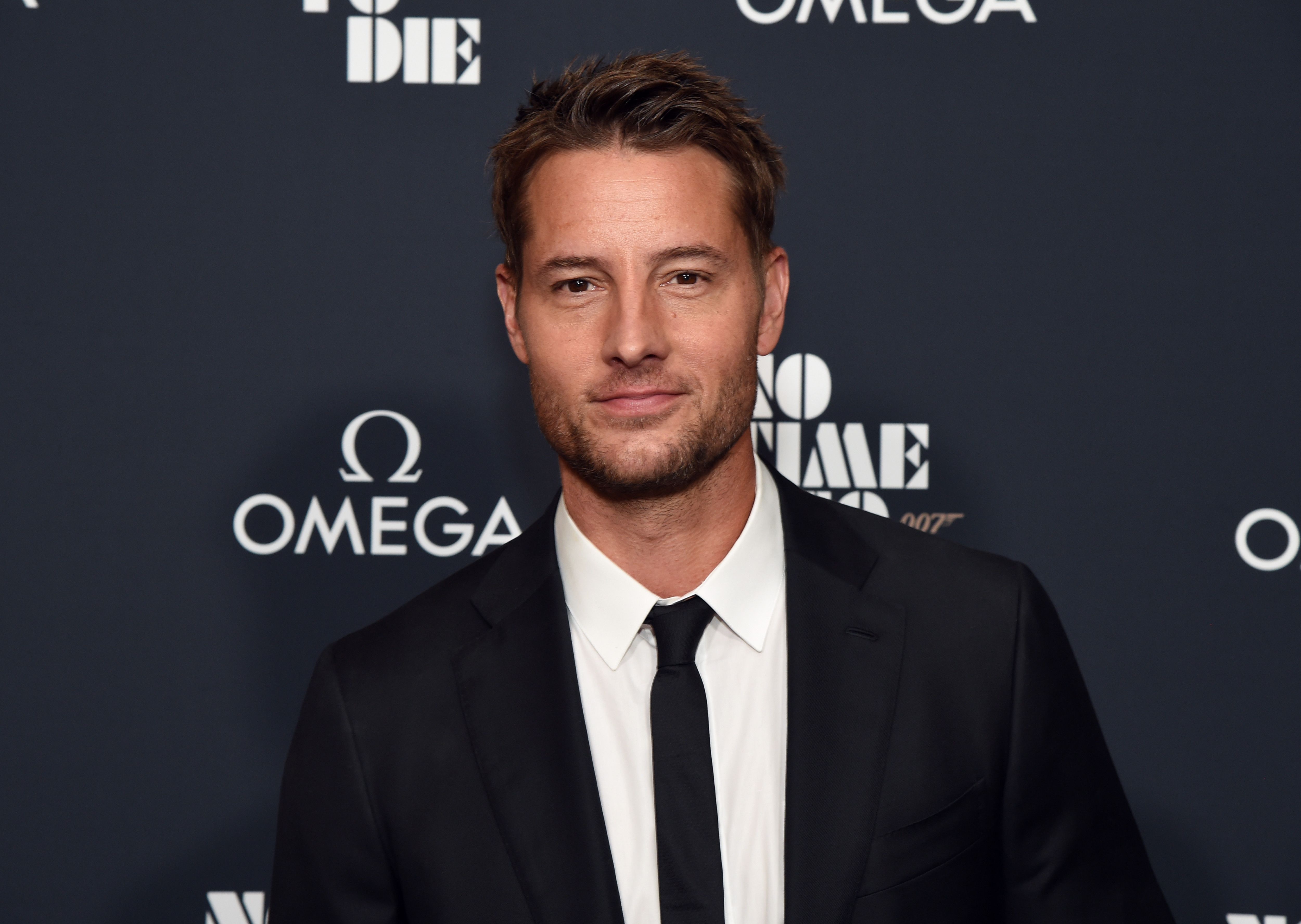 'This Is Us' Season 6 star Justin Hartley wears a black suit over a white button-up shirt and black tie.