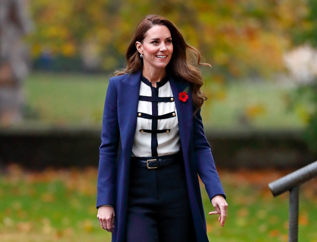 Kate Middleton smiles as she looks on wearing a blue coat, black-and-white shirt, and black pants.