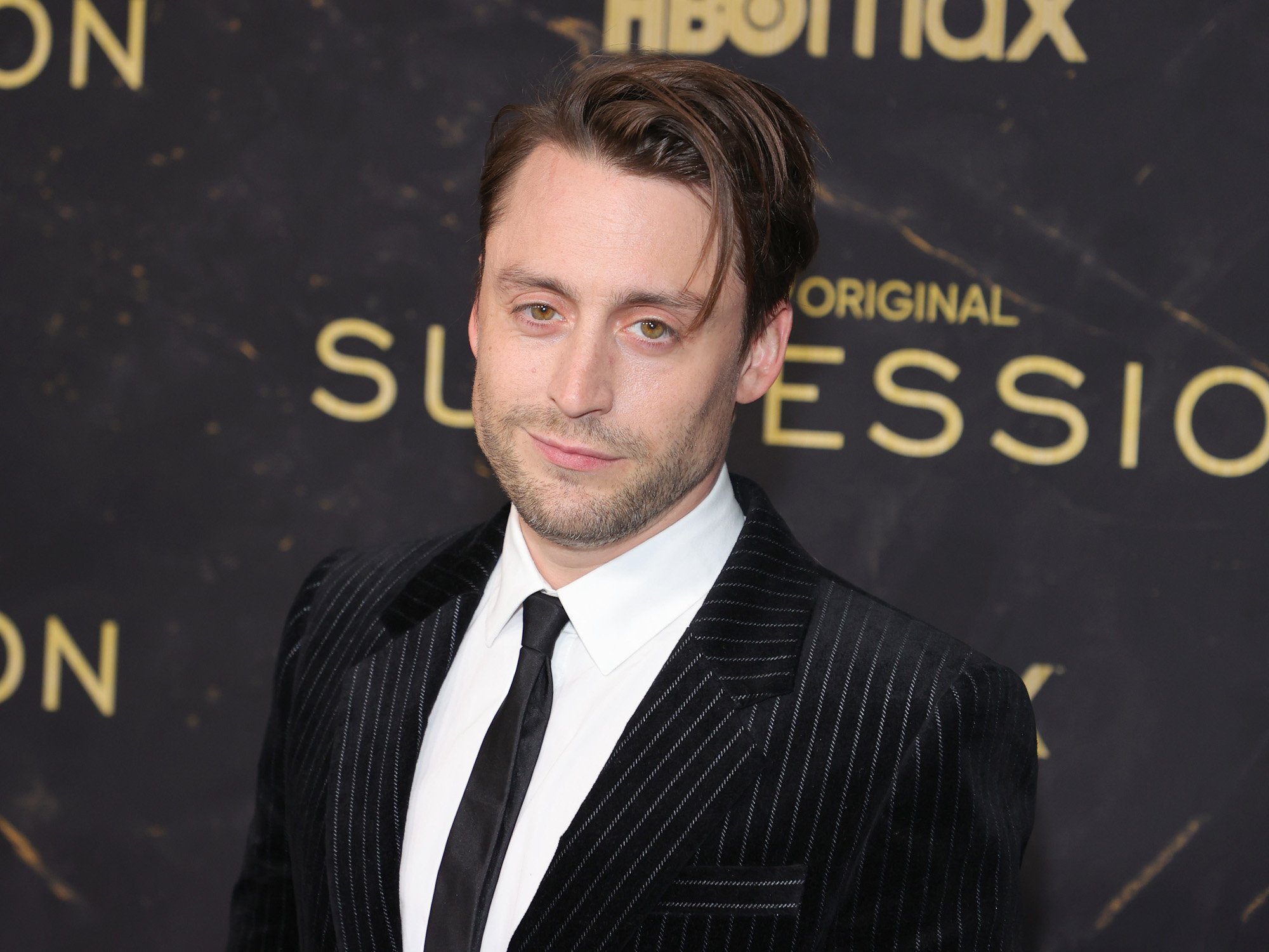 'Succession' star Kieran Culkin, who plays Roman Roy on the HBO show. He's wearing a white shirt and black suit and standing in front of a 'Succession' wall.