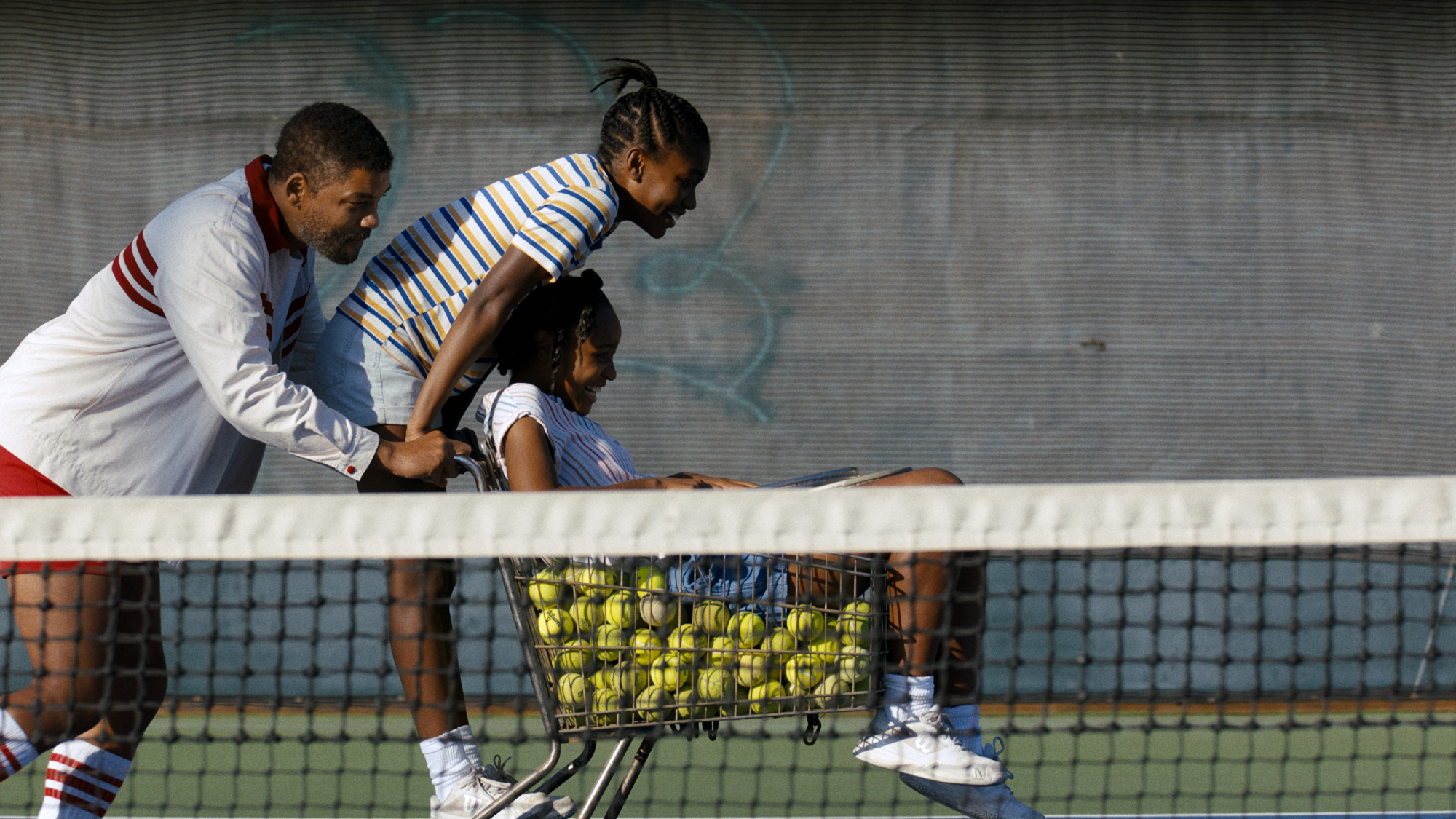'King Richard' review starring Will Smith as Richard Williams, Demi Singleton as Serena Williams, and Saniyya Sidney as Venus Williams with Richard pushing the girls in a tennis ball-filled cart behind the tennis net