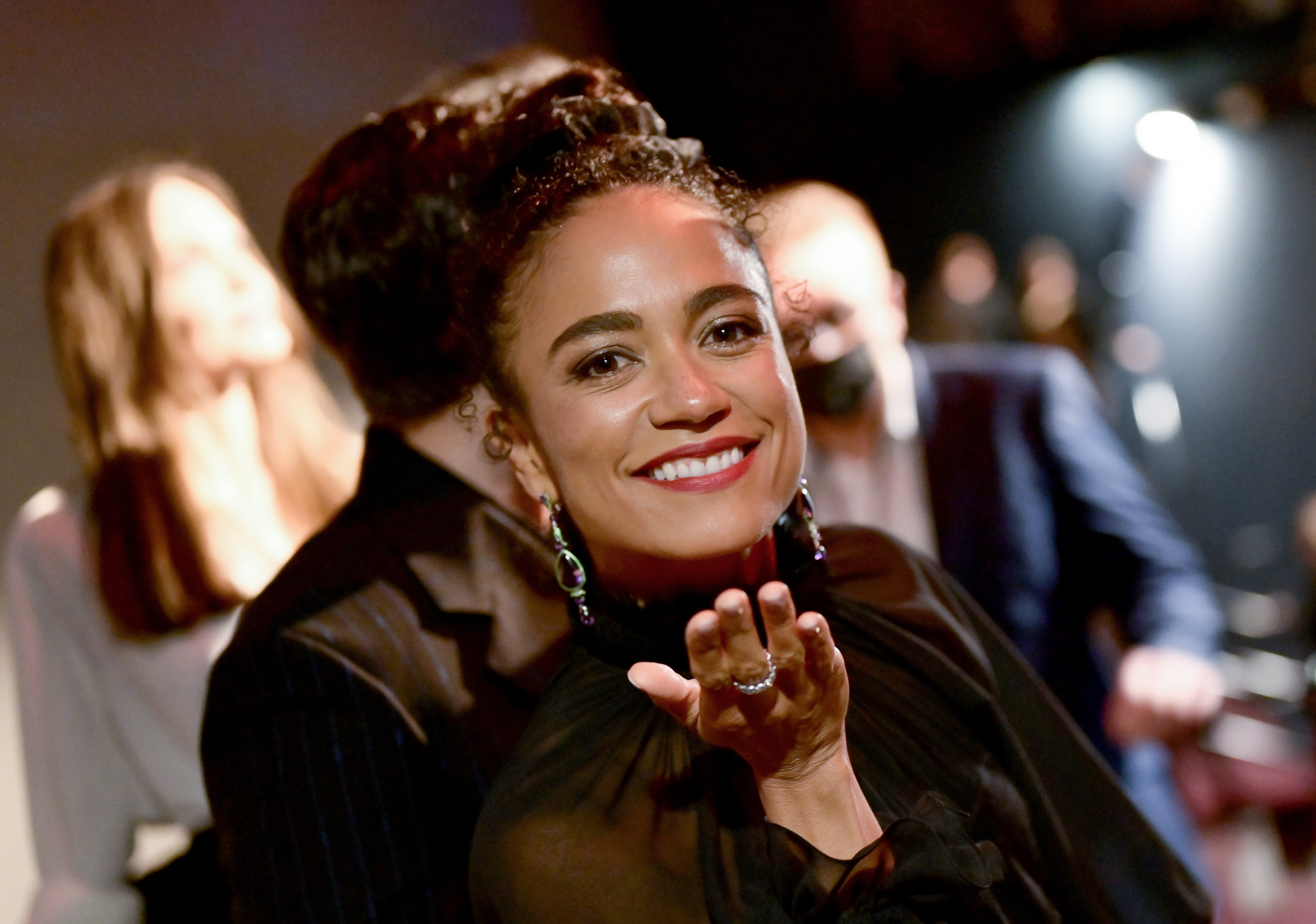 Marvel's 'Eternals' cast member Lauren Ridloff blows a kiss to the camera while wearing a long-sleeved black dress and dangly earrings.