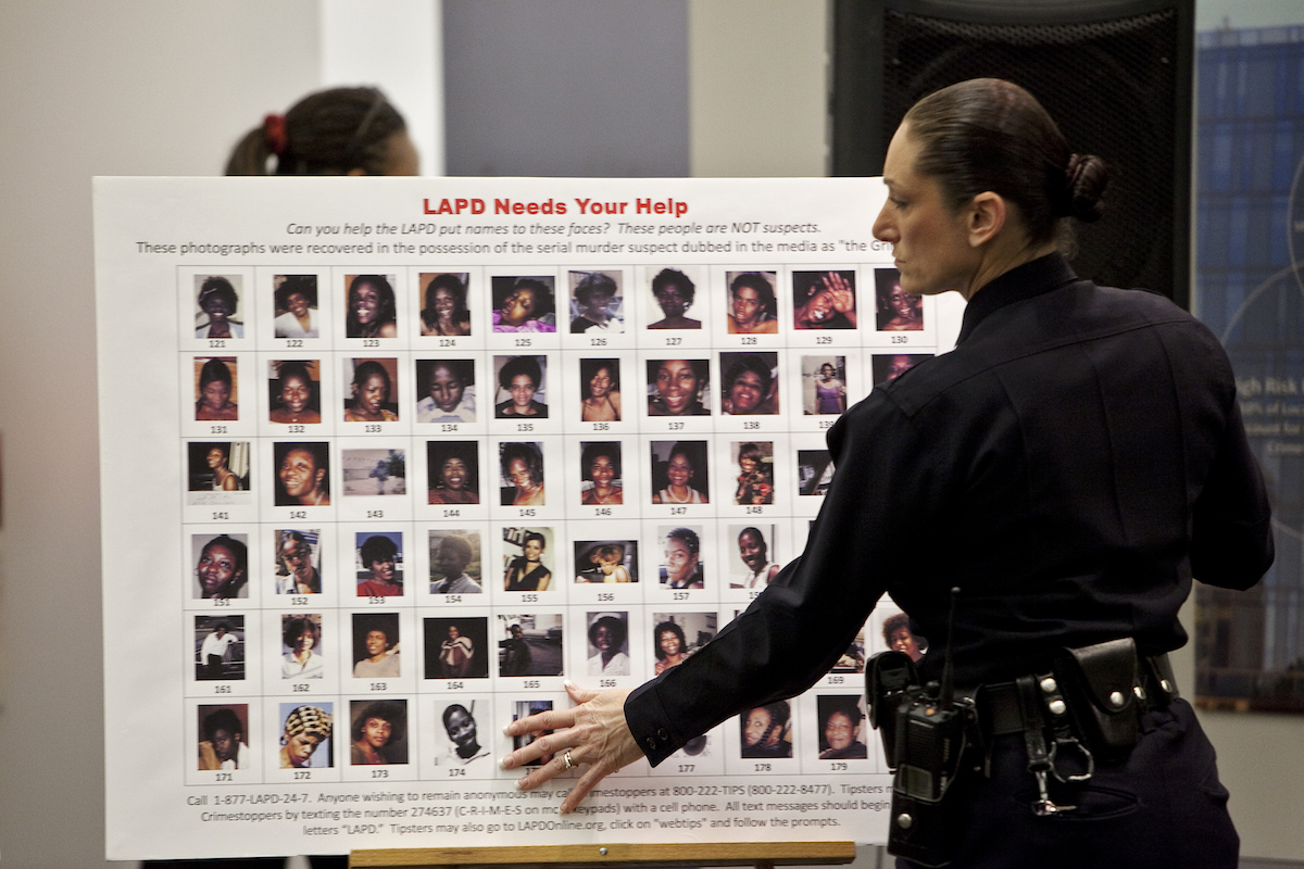 A police officer stands in front of a poster featuring images of women found in possession of killer Lonnie Franklin
