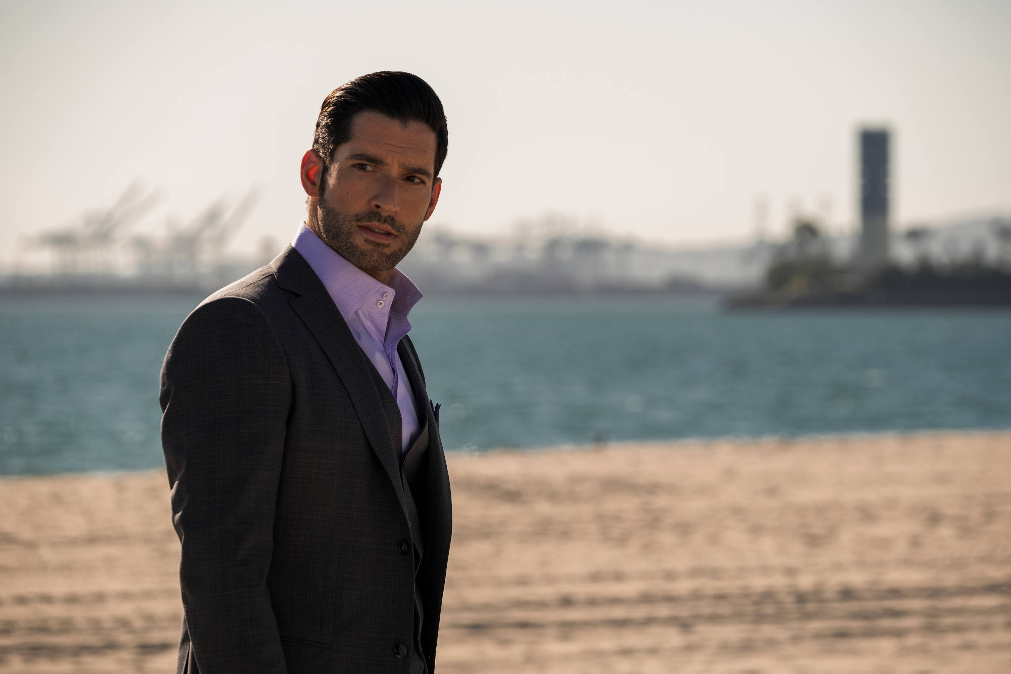 Tom Ellis as Lucifer Morningstar in 'Lucifer' Season 5 on Netflix. He's wearing a suit and standing on the beach.