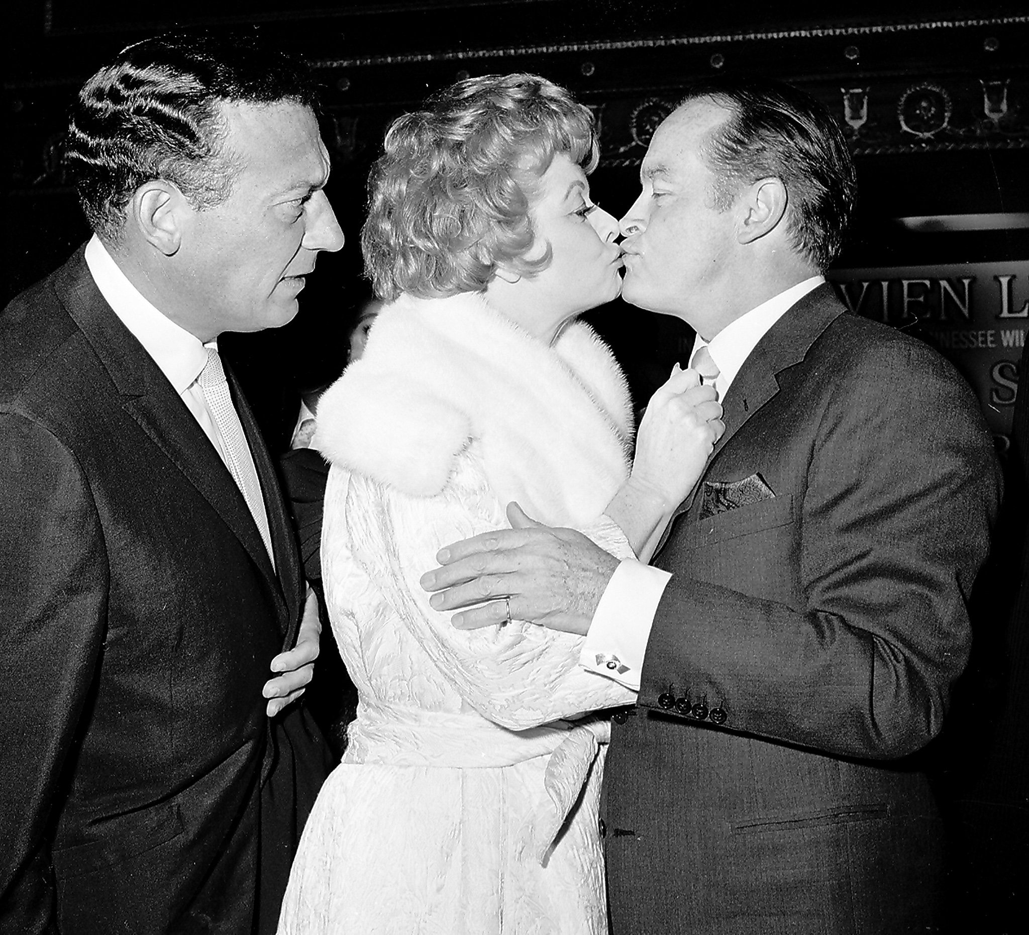 From left to right: Comedian Gary Morton, his wife Lucille Ball, and entertainer Bob Hope. Morton looks on with feigned worry as Ball and Hope greet one another with a kiss.