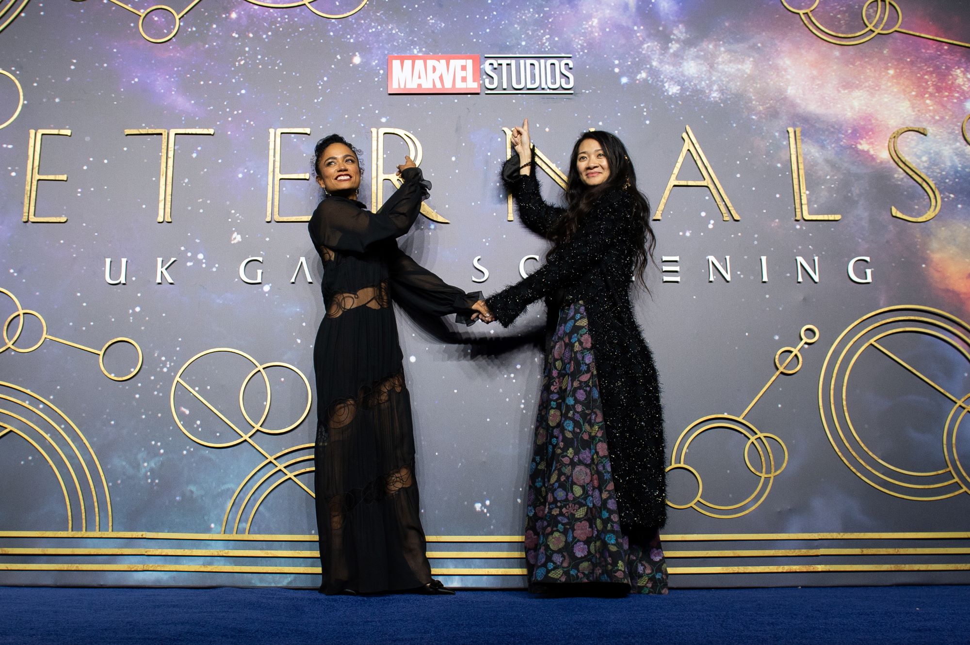 Marvel's 'Eternals' star Lauren Ridloff and director Chloé Zhao point at the wall behind them that says 'Marvel Studios Eternals UK Gala Screening,' and they hold hands. Ridloff wears a black dress with long sleeves. Zhao wears a floral dress with a long black cardigan. Will 'Eternals' get a sequel?
