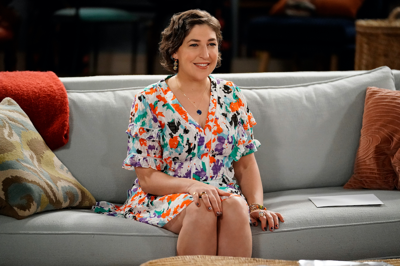 Mayim Bialik smiling in a floral dress and sitting on a couch in a scene from 'Call Me Kat'