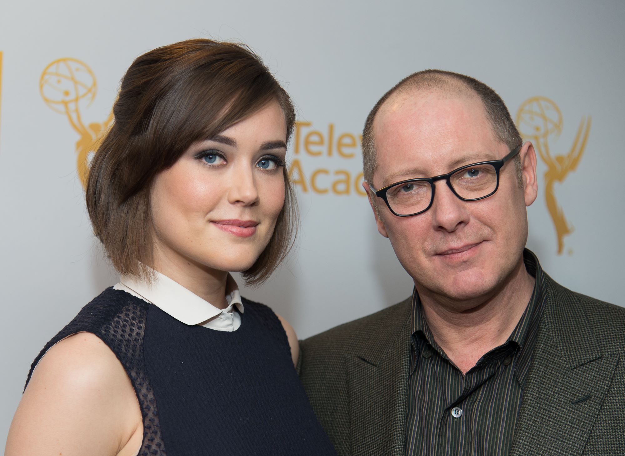 'The Blacklist' stars Megan Boone and James Spader pose together for a picture. Boone wears a blue dress with a white collar. Spader wears a grayish green suit jacket over a green and gray striped button-up shirt.