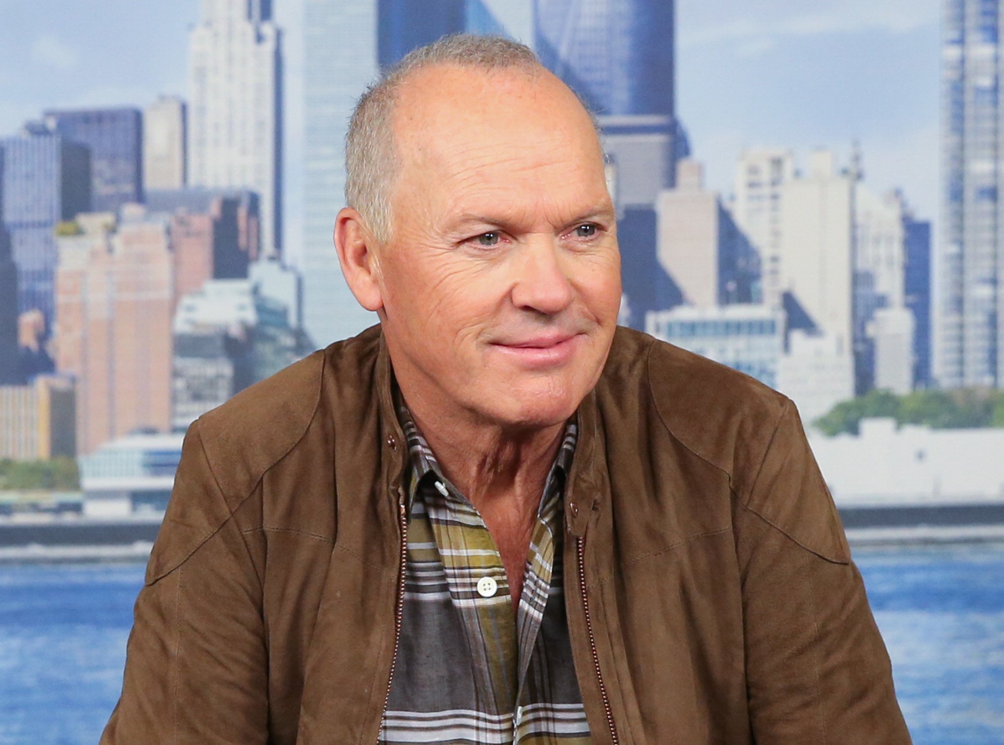 Michael Keaton promoting 'Spider-Man: Homecoming,' where he plays the Spider-Man villain Vulture. He's wearing a plaid shirt and brown jacket, and the image of New York City is behind him.