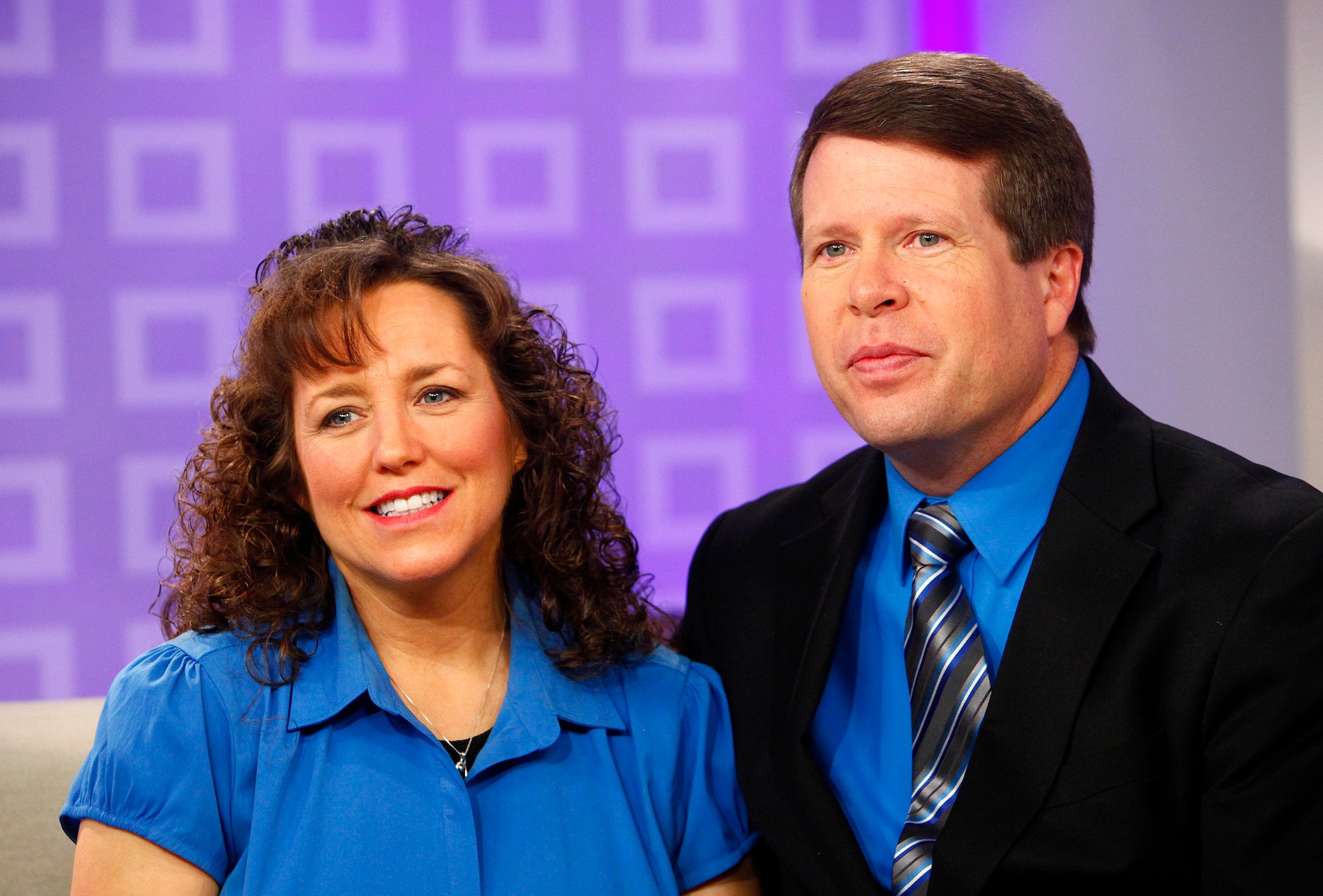 Michelle Duggar and Jim Bob Duggar sitting down against a purple background