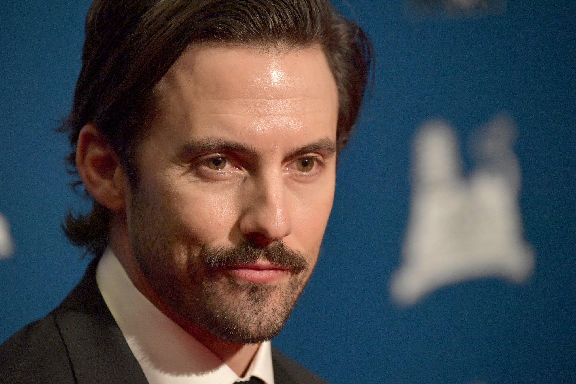 'Gilmore Girls' star Milo Ventimiglia, who played Rory's boyfriend Jess on the show. He's wearing a suit and standing in front of a blue wall.