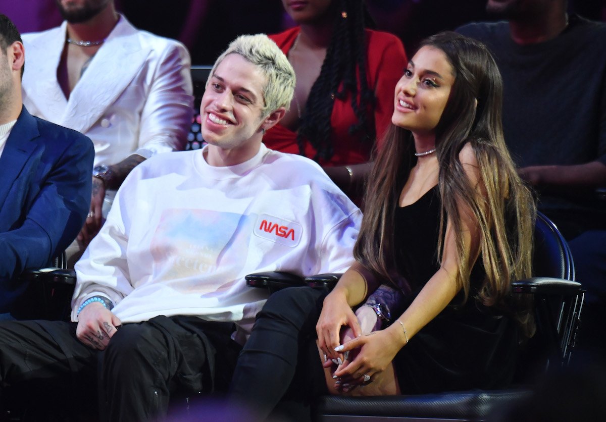 Pete Davidson and Ariana Grande sit together and hold hands at an event.