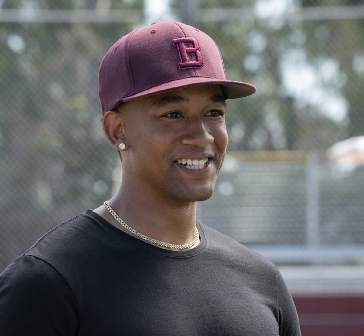 Peyton Alex Smith wearing a burgundy hat and black shirt on a baseball field in the 'All American' spinoff series.