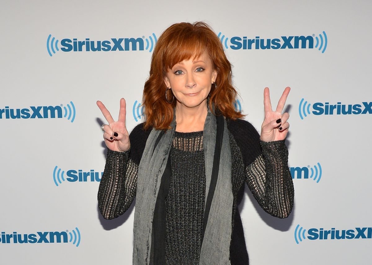 Reba McEntire in a black sweater and gray scarf, flashing peace signs with both hands