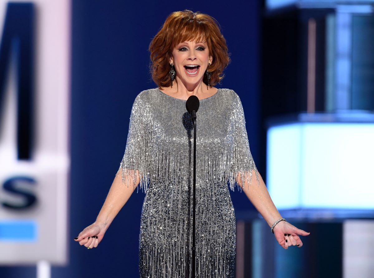 Reba McEntire in a silver dress, extending her arms out to each side and speaking into a microphone