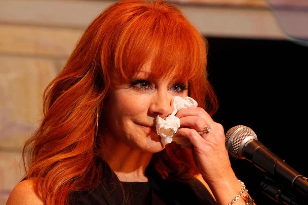 Reba McEntire seated in front of a microphone, dabbing her face with a tissue