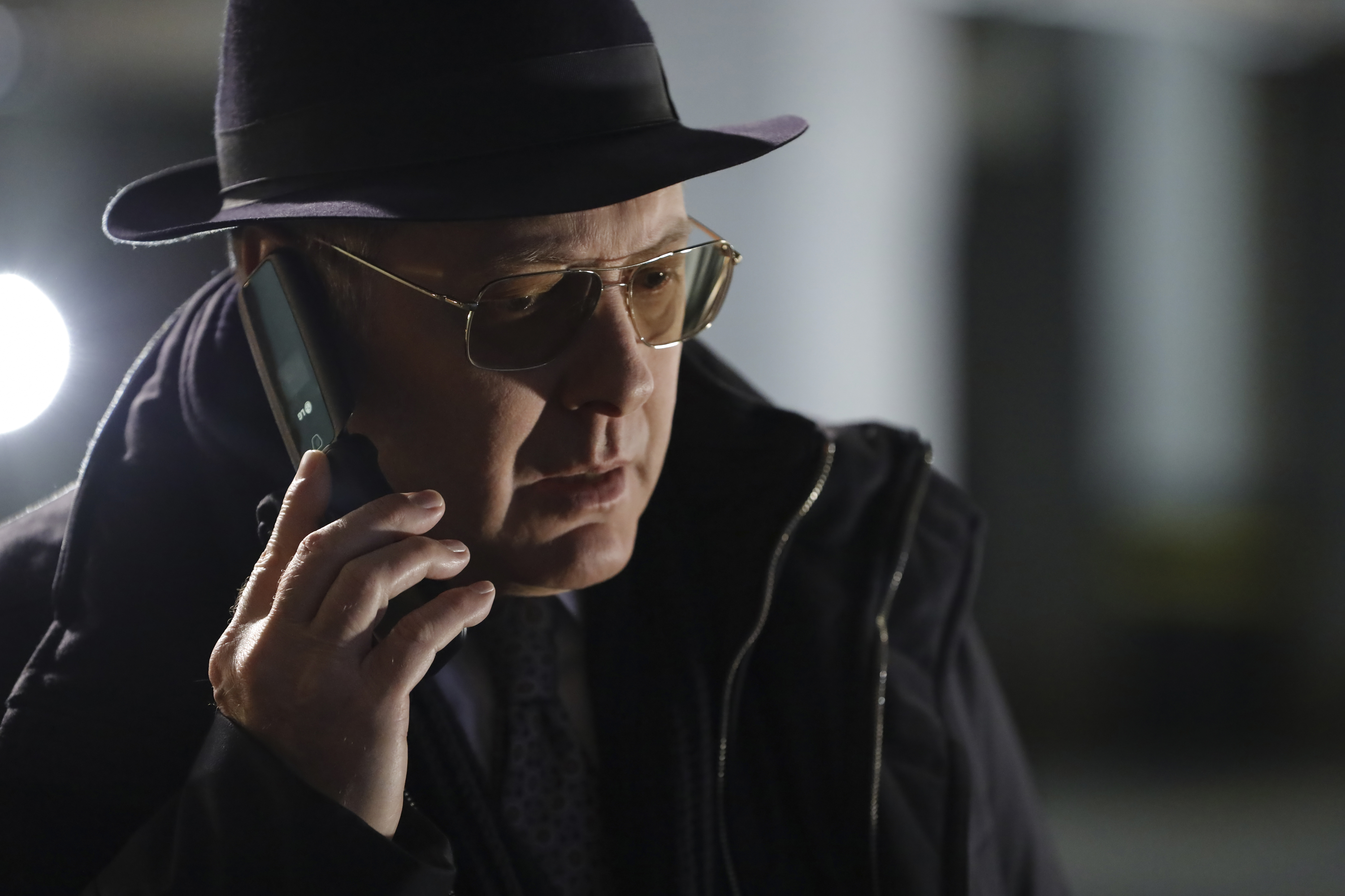 'The Blacklist' star James Spader, in character as Red, subject of the Redarina fan theory, wears a black winter jacket over a suit and tie, and a pair of tinted glasses and a black hat. He holds his flip phone to his ear.
