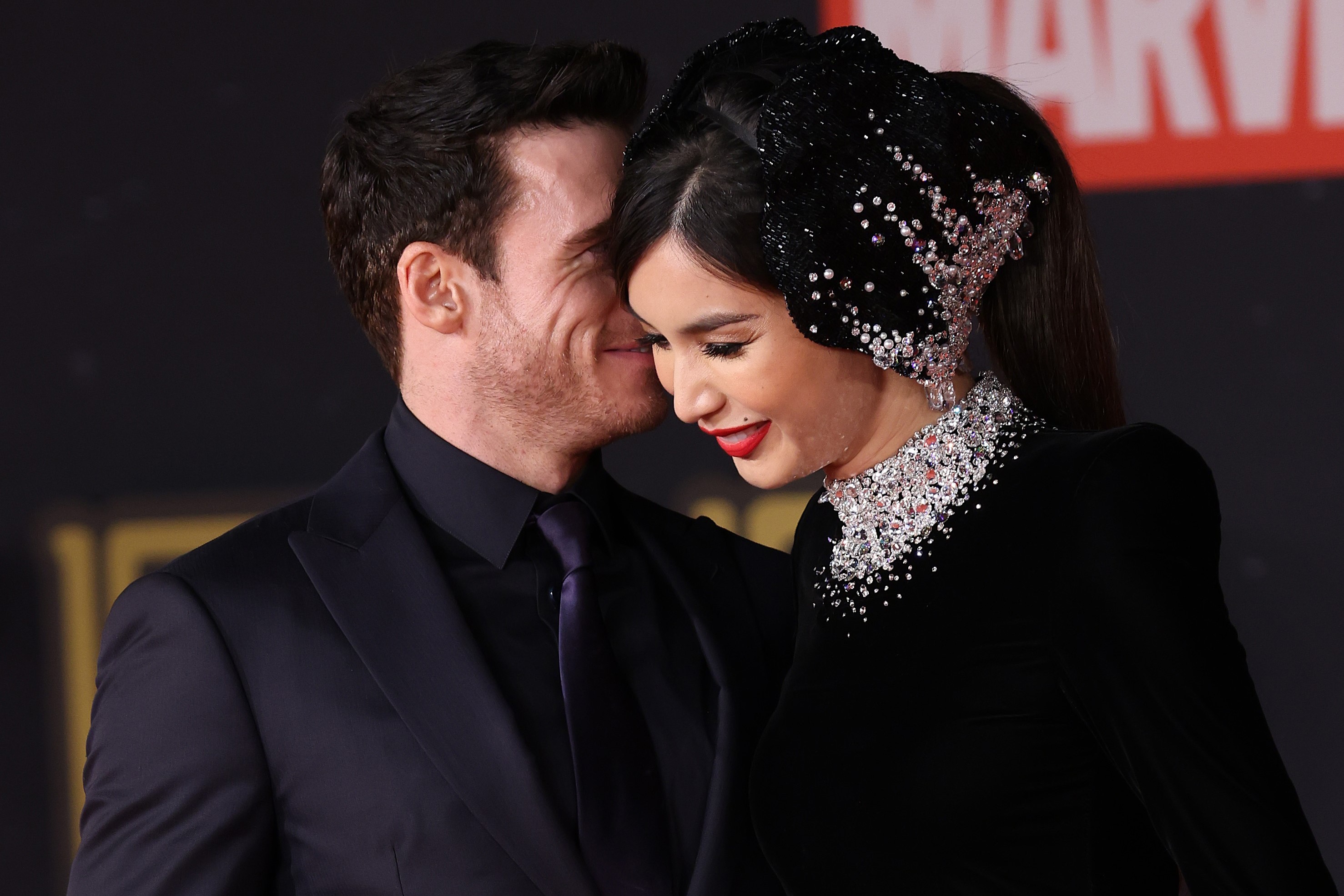 'Eternals' stars Richard Madden and Gemma Chan on the red carpet. Madden wears a dark blue suit and tie. Chan wears a black dress that is studded with diamonds along the collar, and she wears a black headpiece studded with diamonds and pearls. Madden is whispering something into Chan's ear, and she is smiling. Their two characters have a sex scene in the film.