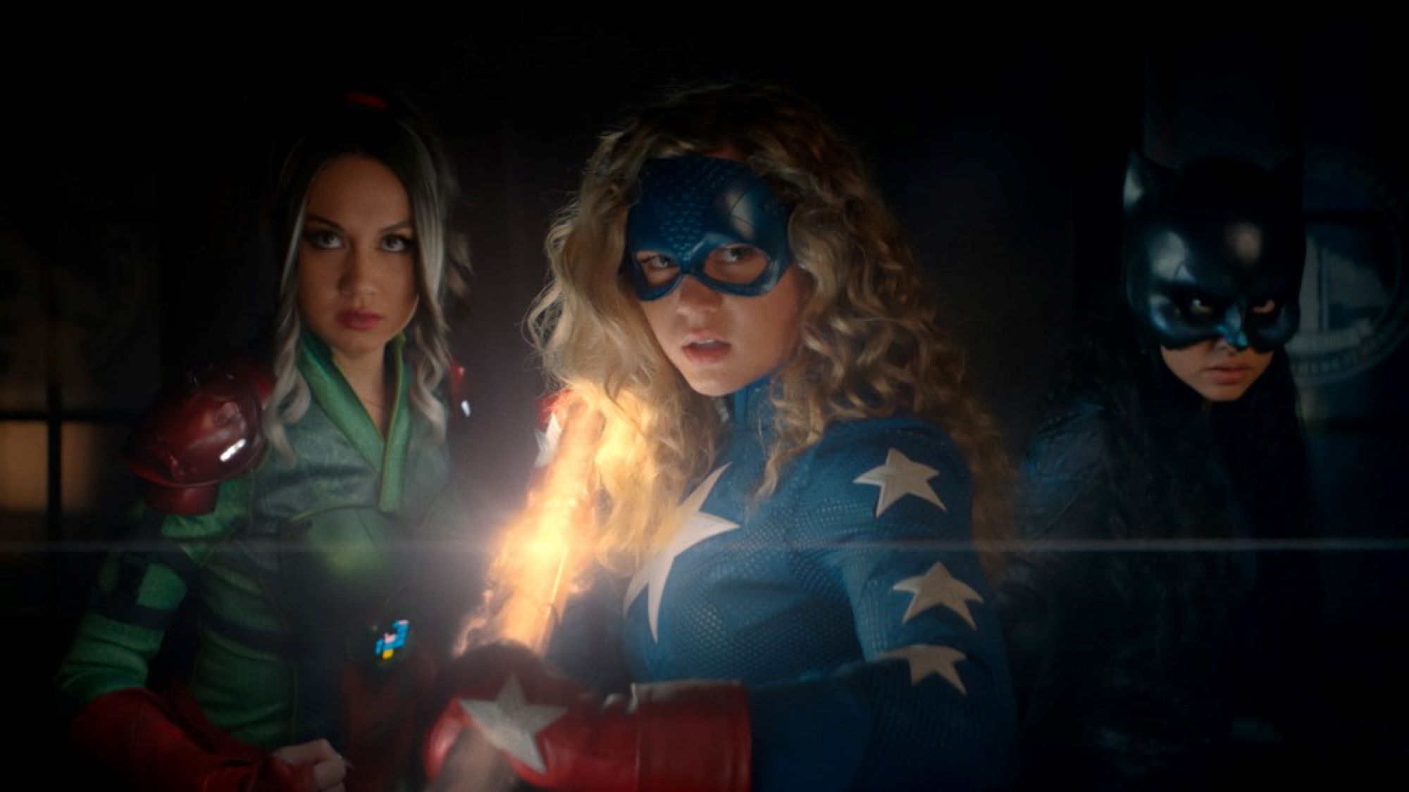 'Stargirl' actors Meg DeLacy, Brec Bassinger, and Yvette Monreal, in character as Cindy, Courtney, and Yolanda, prepare for a fight. DeLacy wears her green and red Shiv costume. Bassinger wears her red, white, and blue Stargirl costume and carries the Cosmic Staff. Monreal wears her black Wildcat costume.