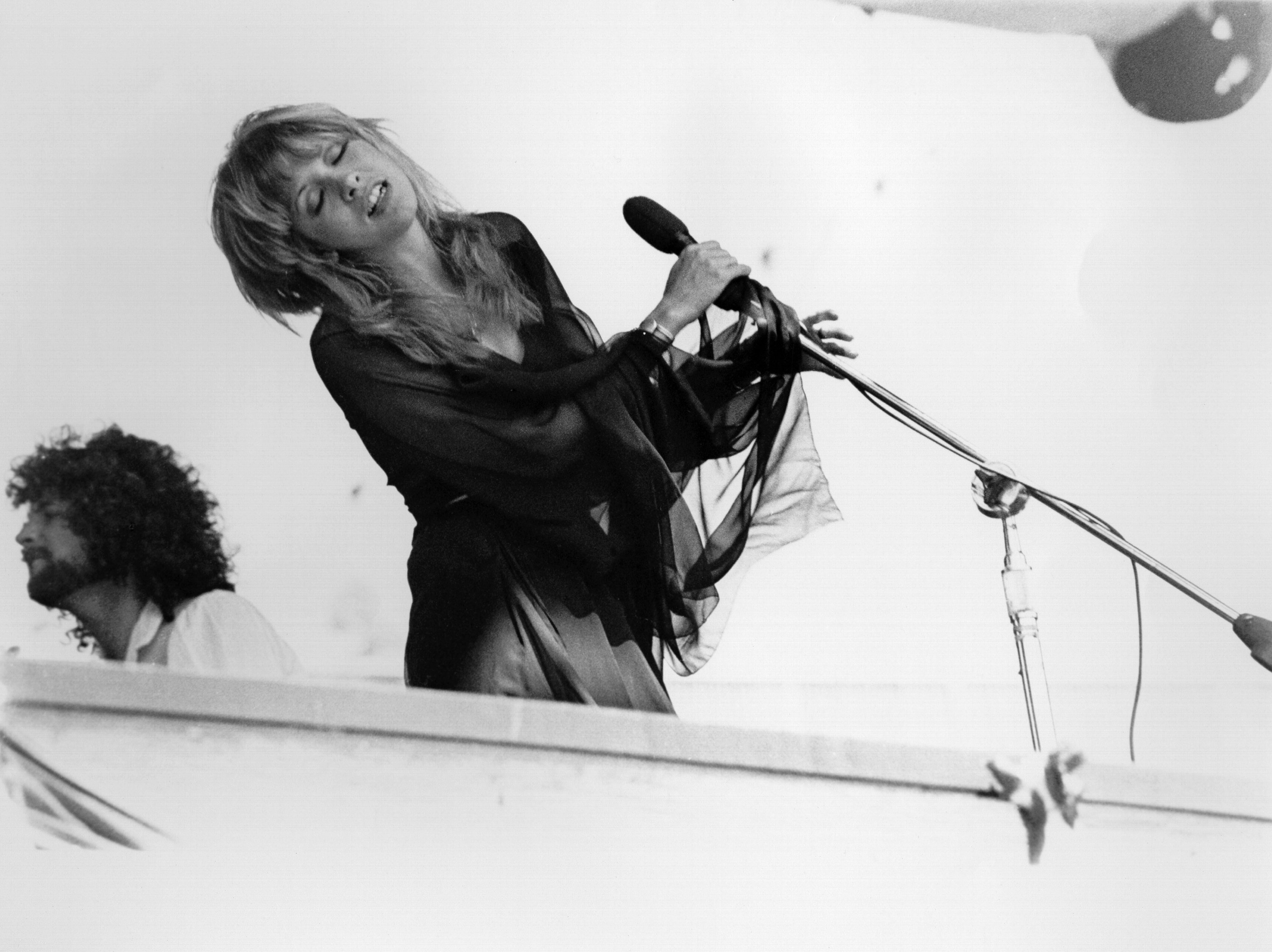 A black and white photo of Stevie Nicks wearing a black chiffon dress. She holds a microphone and has her eyes closed.