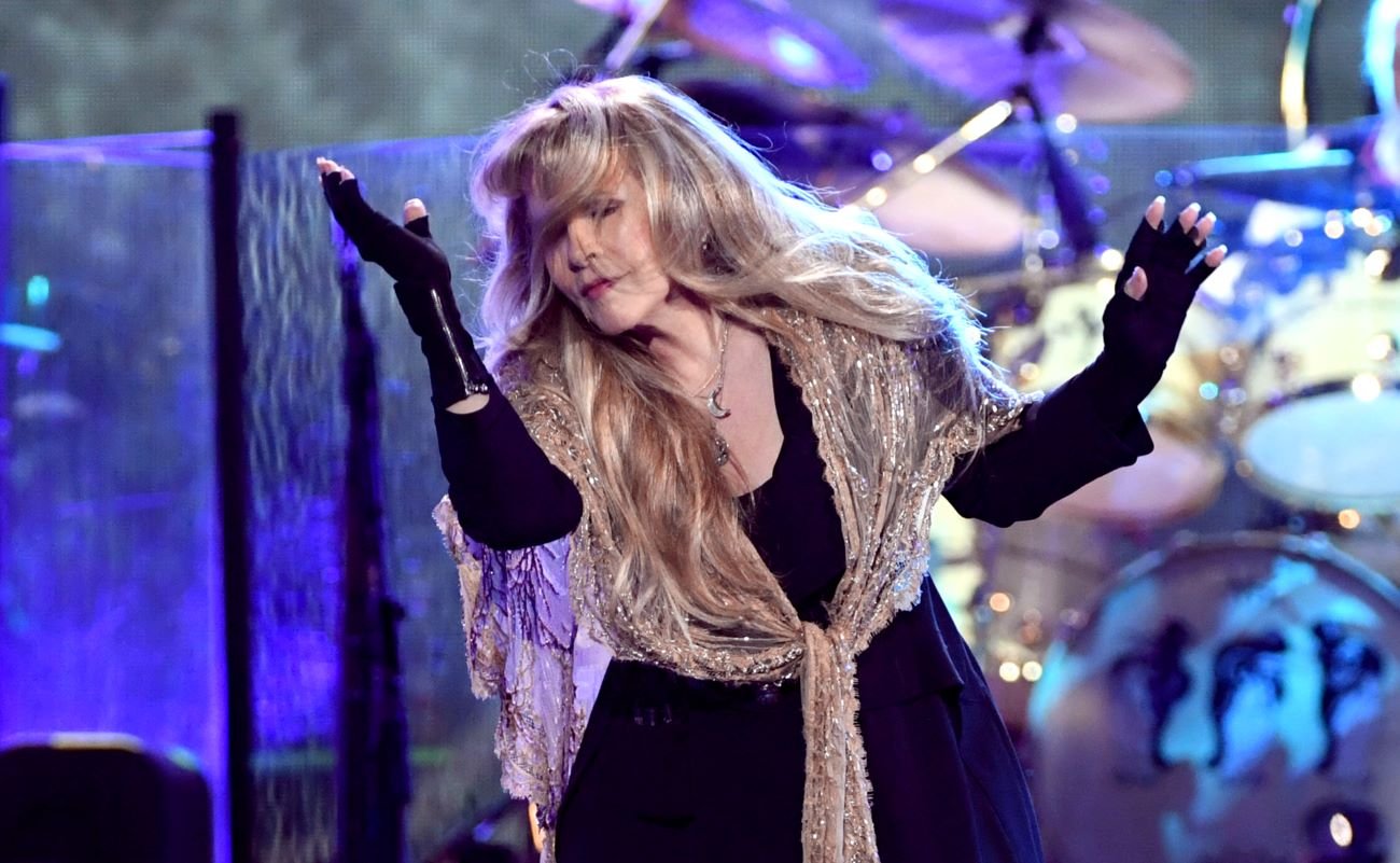 Stevie Nicks wears a black dress, black fingerless gloves, and a tan shawl. She dances onstage. 