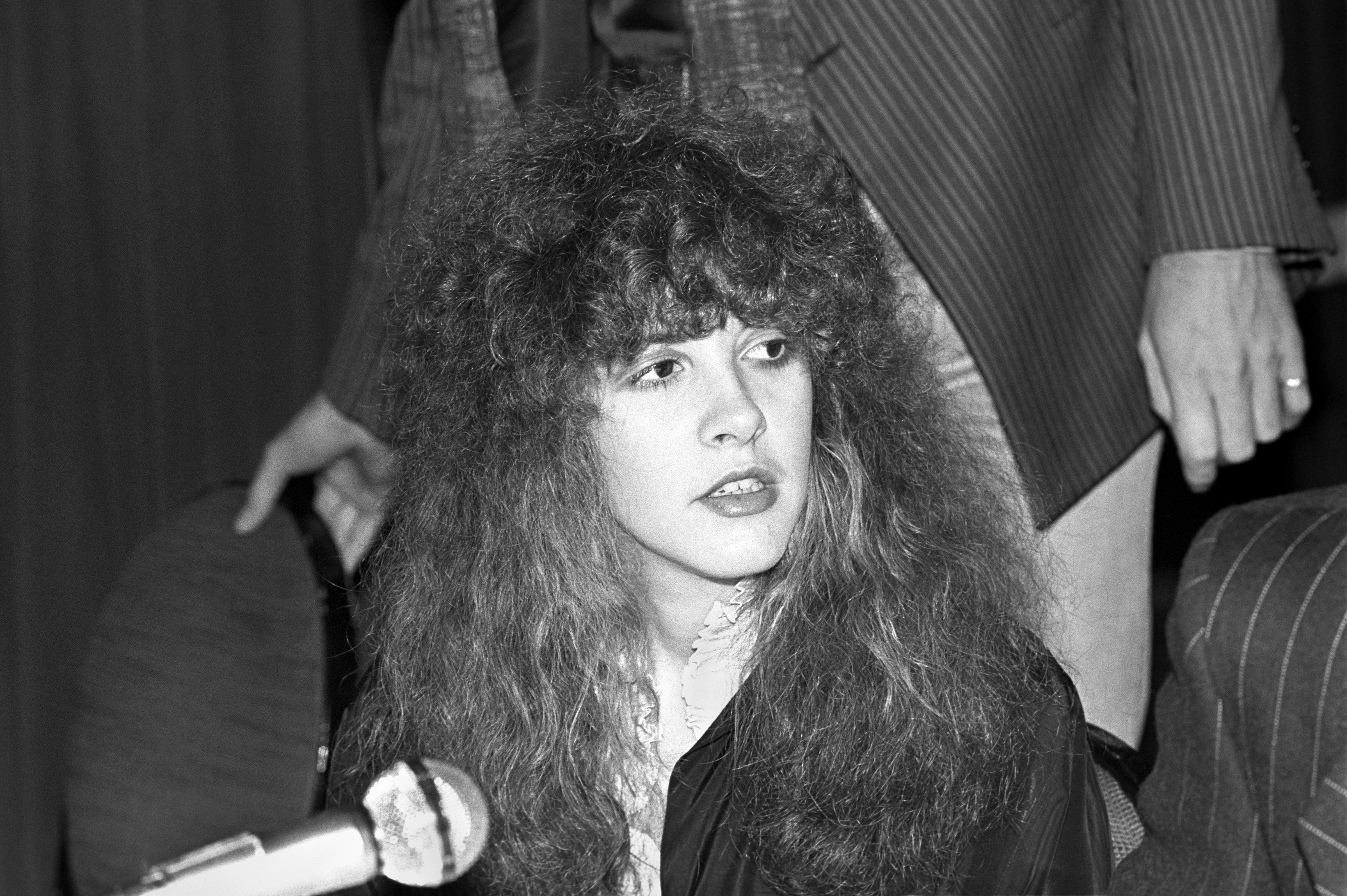 A black and white picture of a young Stevie Nicks wearing a black sweater and white shirt. She sits in front of a microphone.