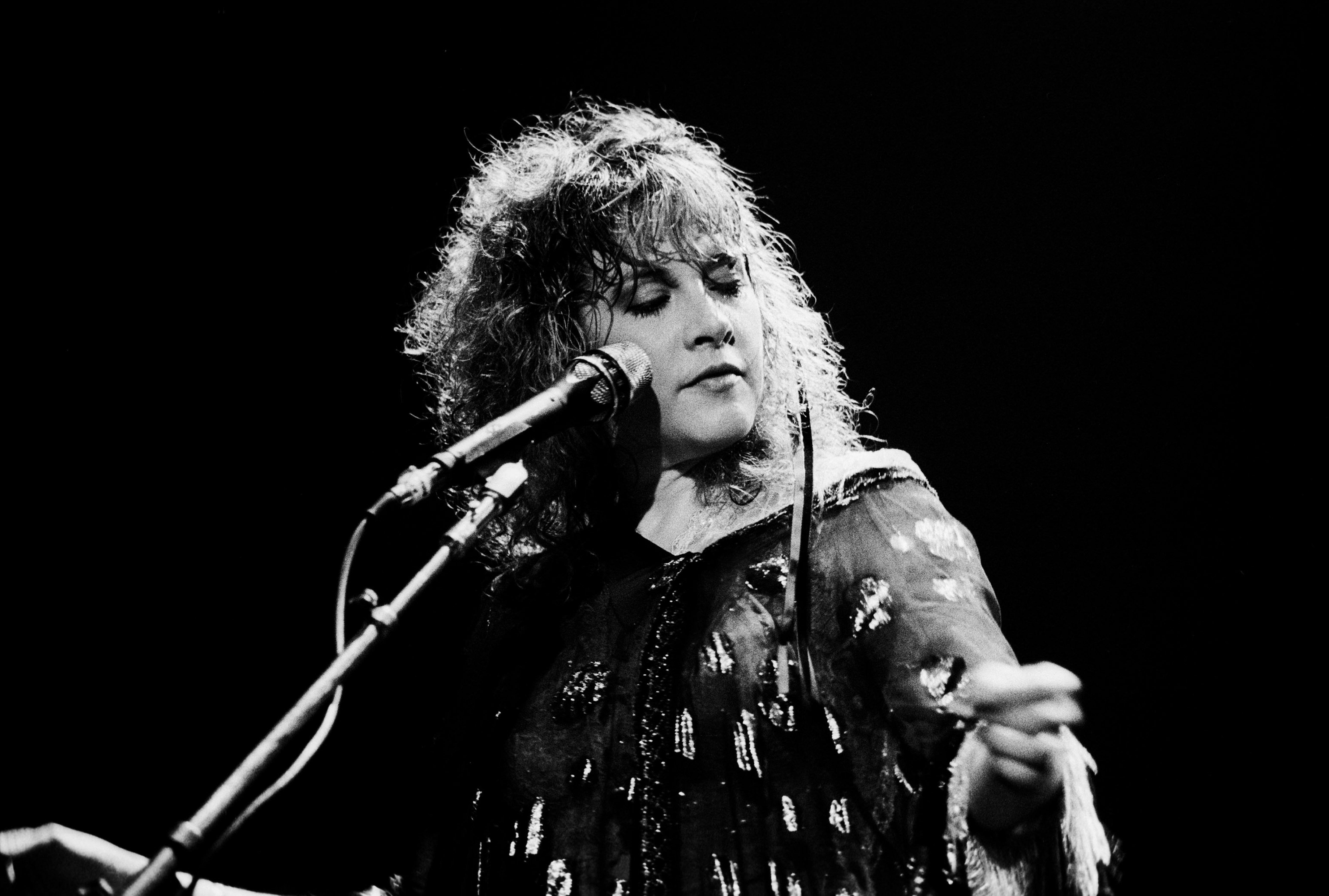 A black and white photo of Stevie Nicks wearing a dress and standing in front of a microphone.