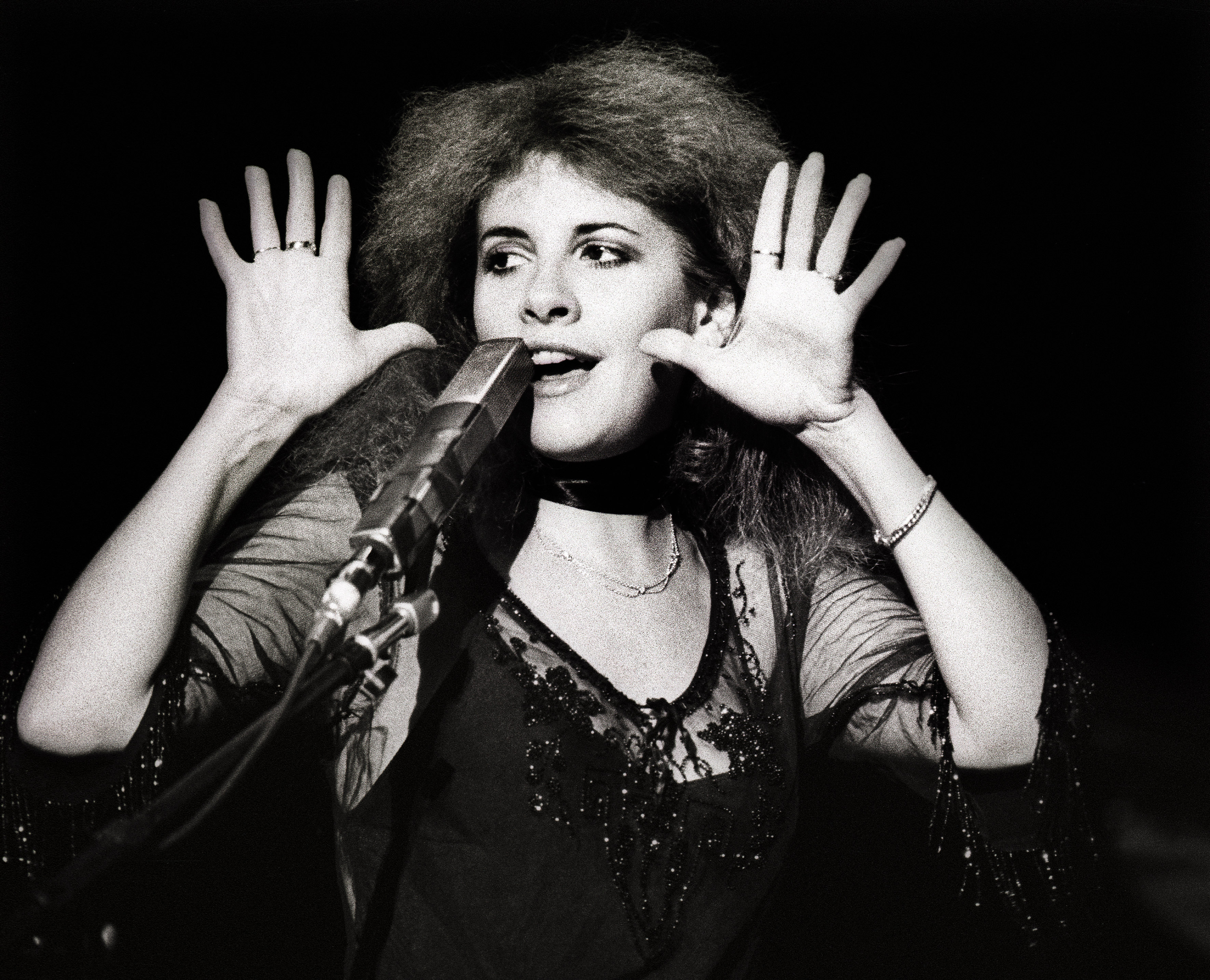 A black and white photo of Fleetwood Mac's Stevie Nicks wearing a lace shirt. She holds her hands up near her face and stands in front of a microphone.