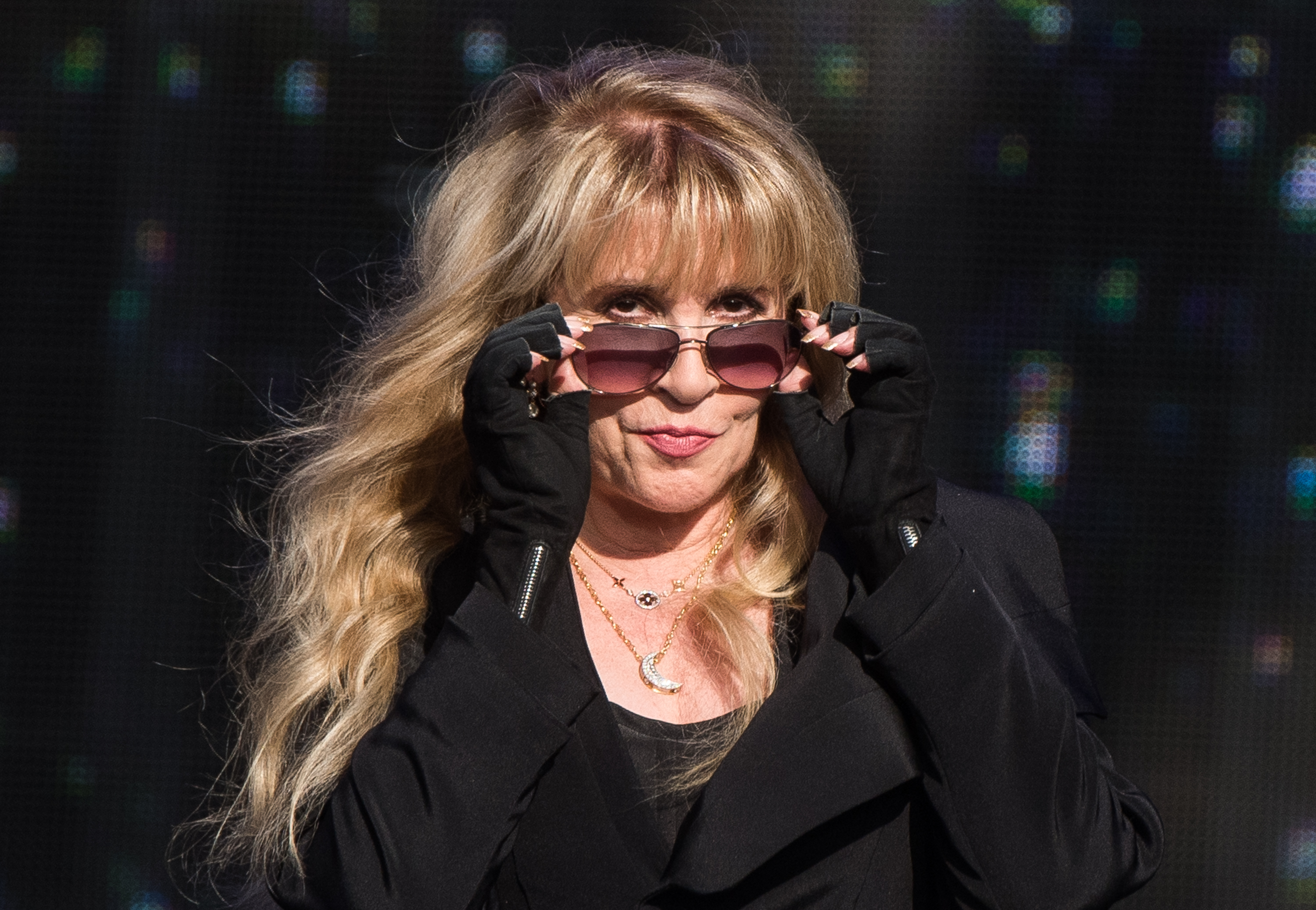 Stevie Nicks wears a black dress, black fingerless gloves, and sunglasses.