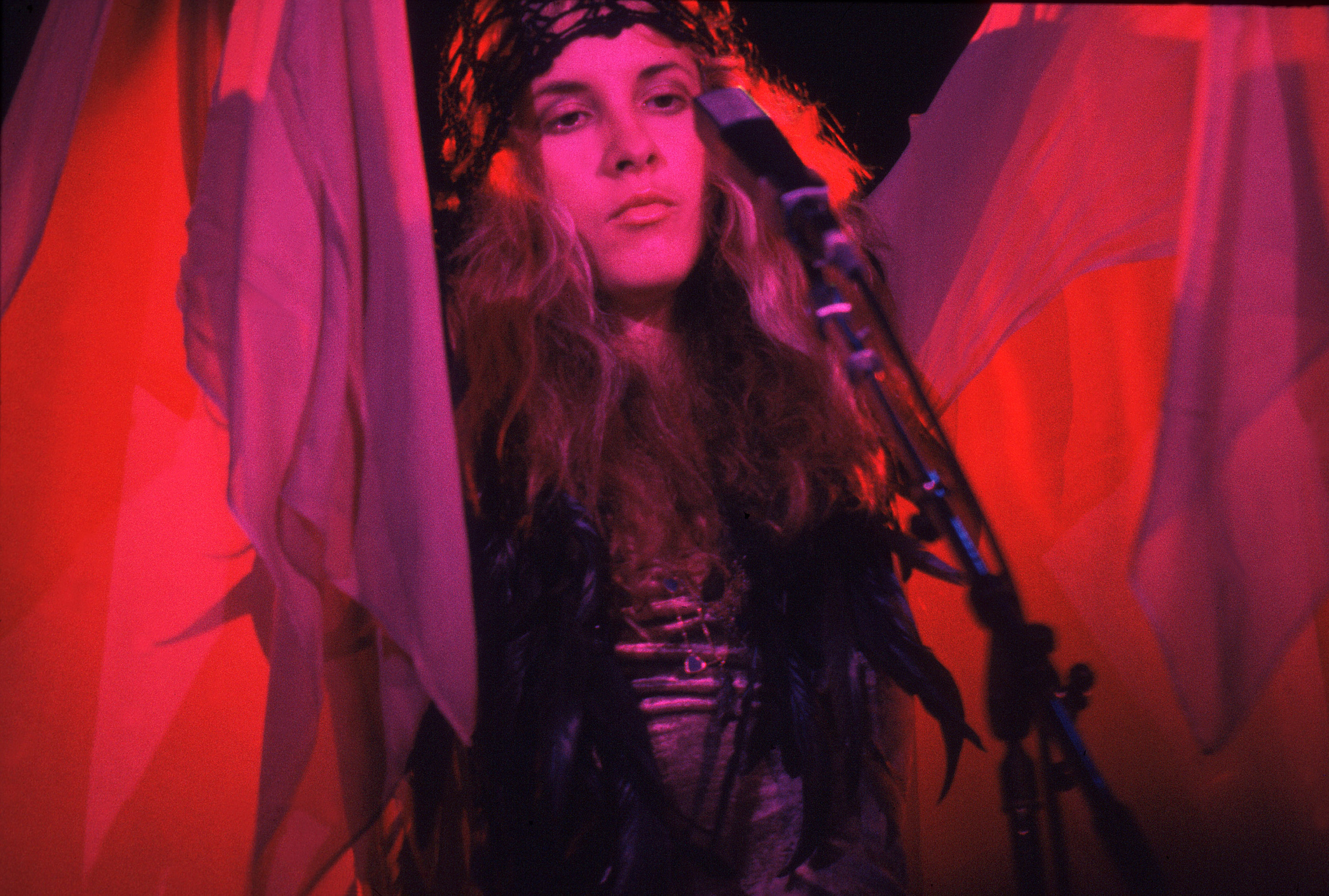 Stevie Nicks wears a scarf over her head and holds her shawl up. She's lit in pink light and stands in front of a microphone.