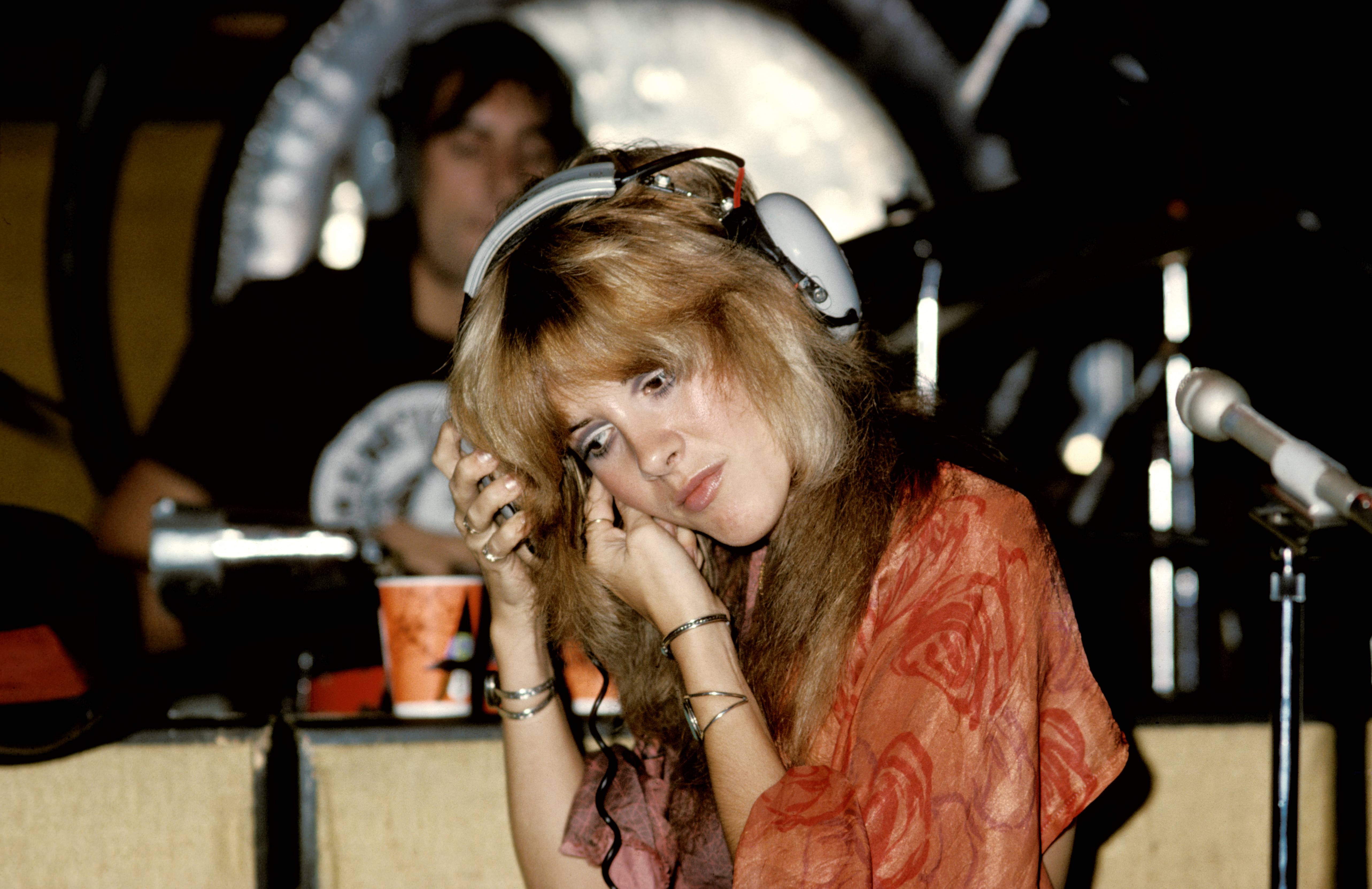 A young Stevie Nicks wears an orange floral shirt and gray headphones while recording with Fleetwood Mac.