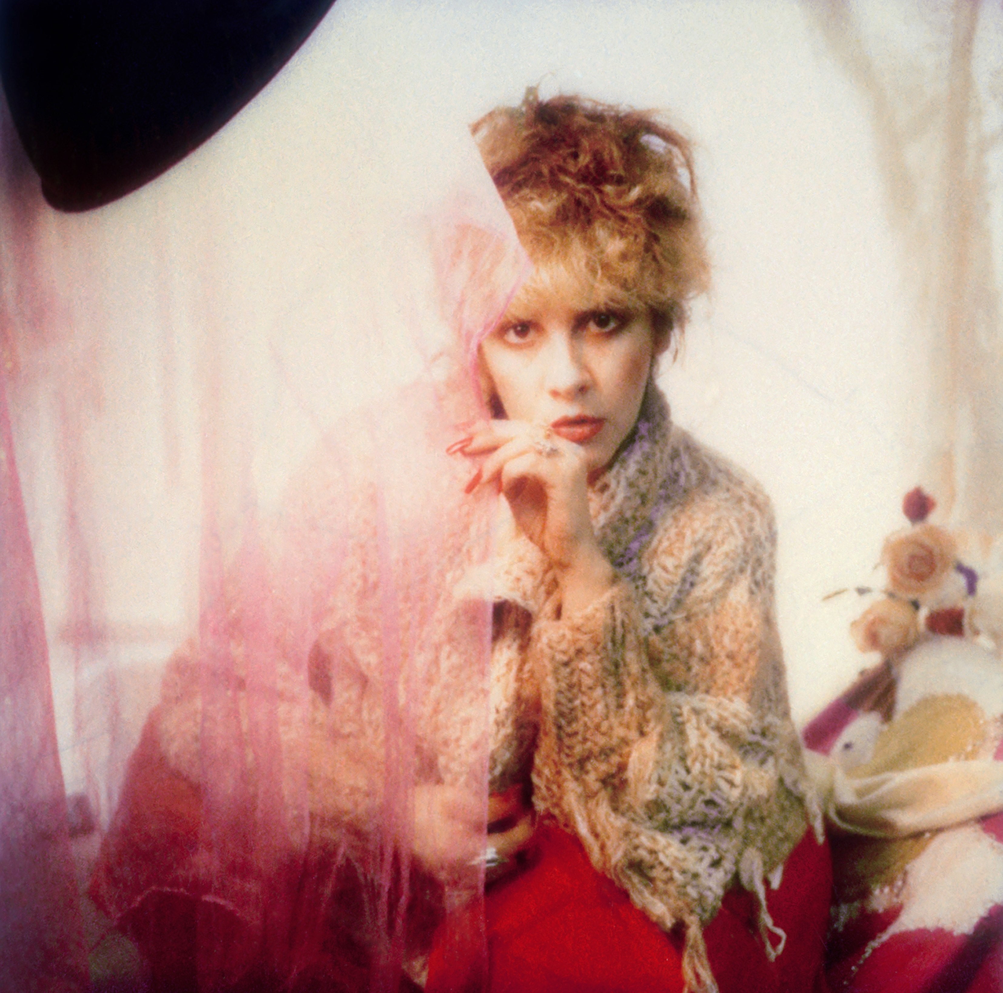 Stevie Nicks wears a knit sweater over a red dress. She looks behind a sheer pink curtain.