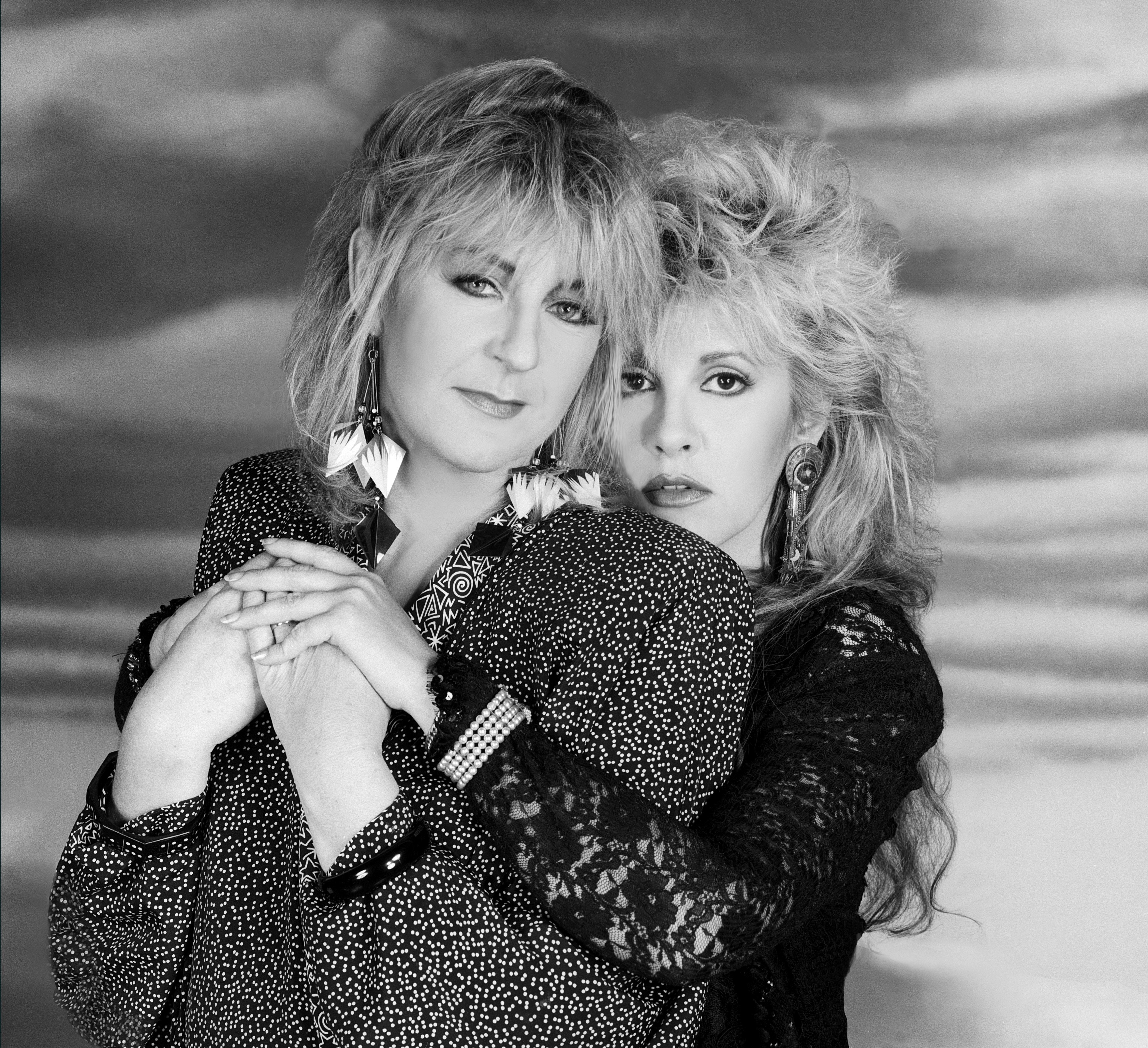 Christine McVie wears a dotted blouse and Stevie Nicks wears a lace shirt. Nicks wraps her arms around McVie.