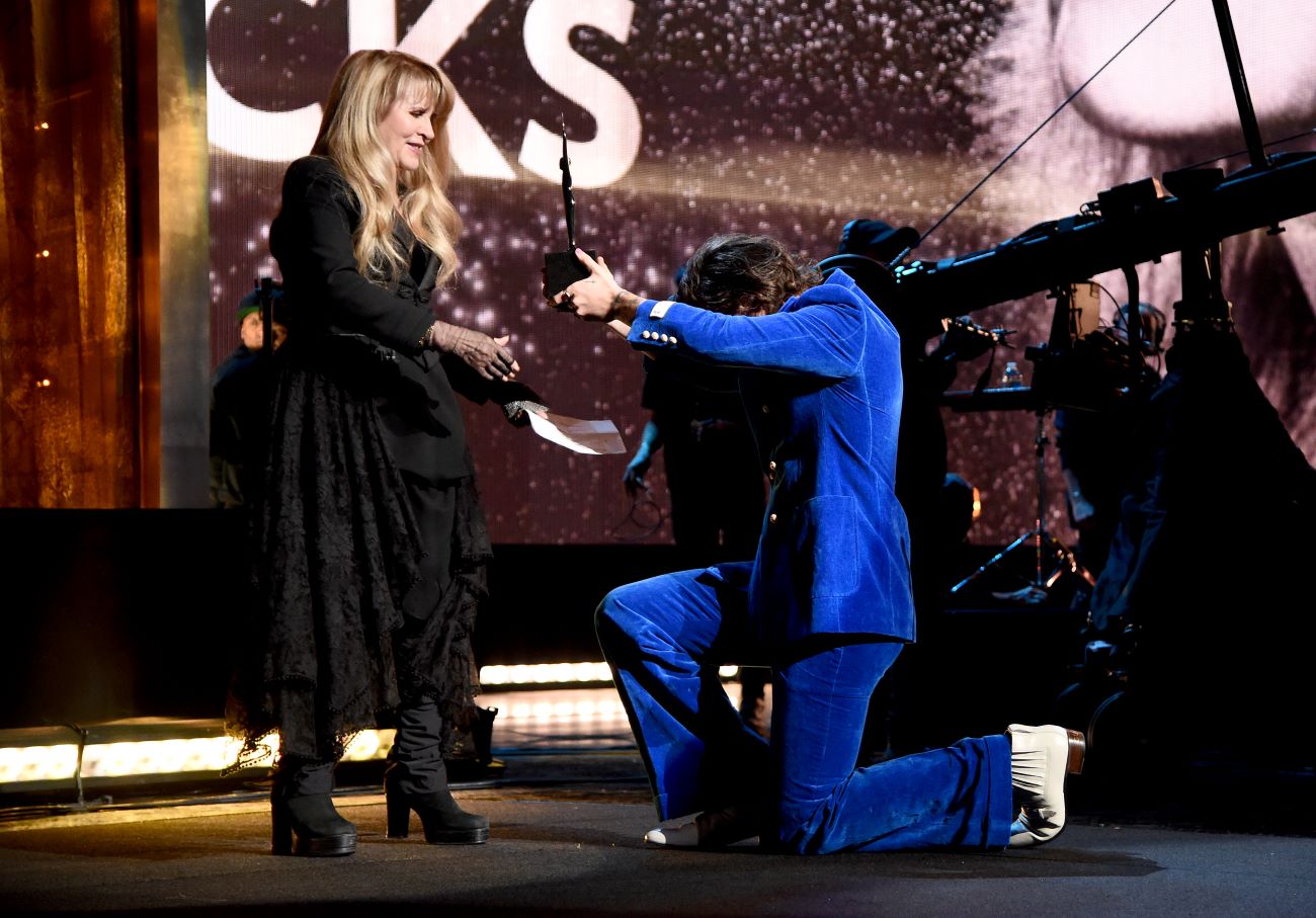 Stevie Nicks wears a black dress and accepts an award from Harry Styles, who wears a blue suit. He kneels in front of her.