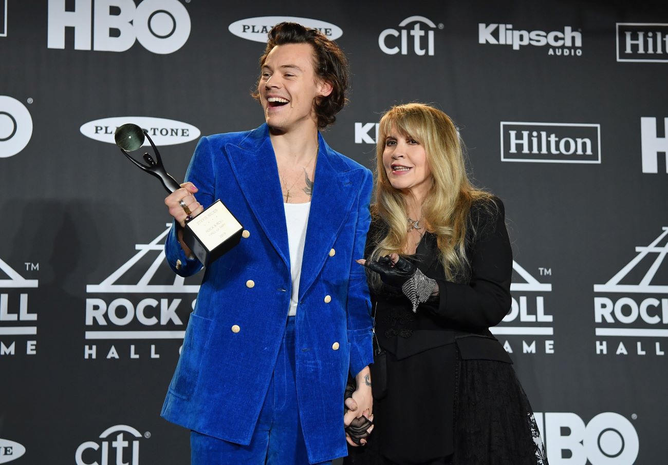 Harry Styles wears a blue suit and holds an award. Stevie Nicks wears a black dress and holds his hand. 