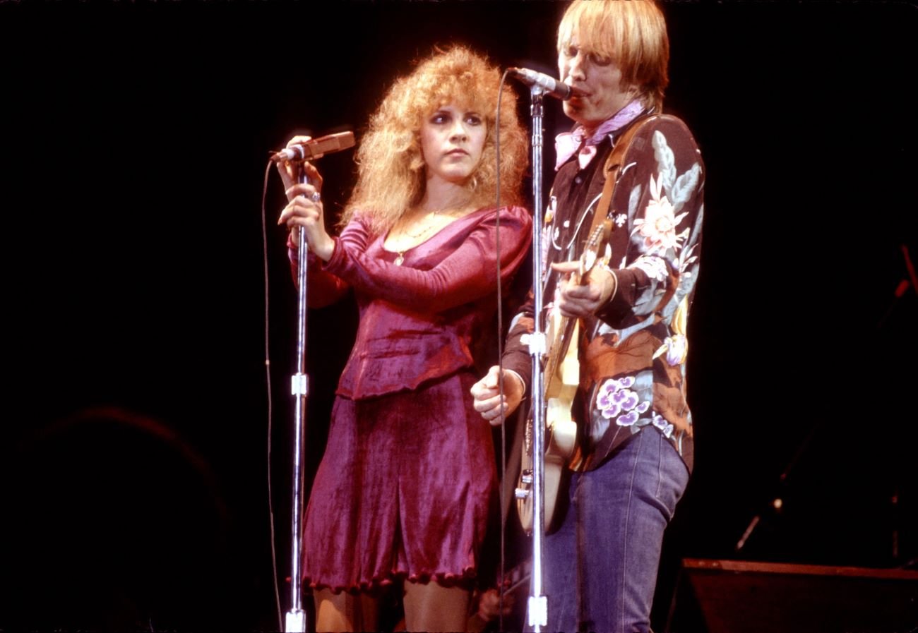 Stevie Nicks wears a purple dress and Tom Petty wears a black floral shirt. He sings and plays guitar and she holds a microphone.