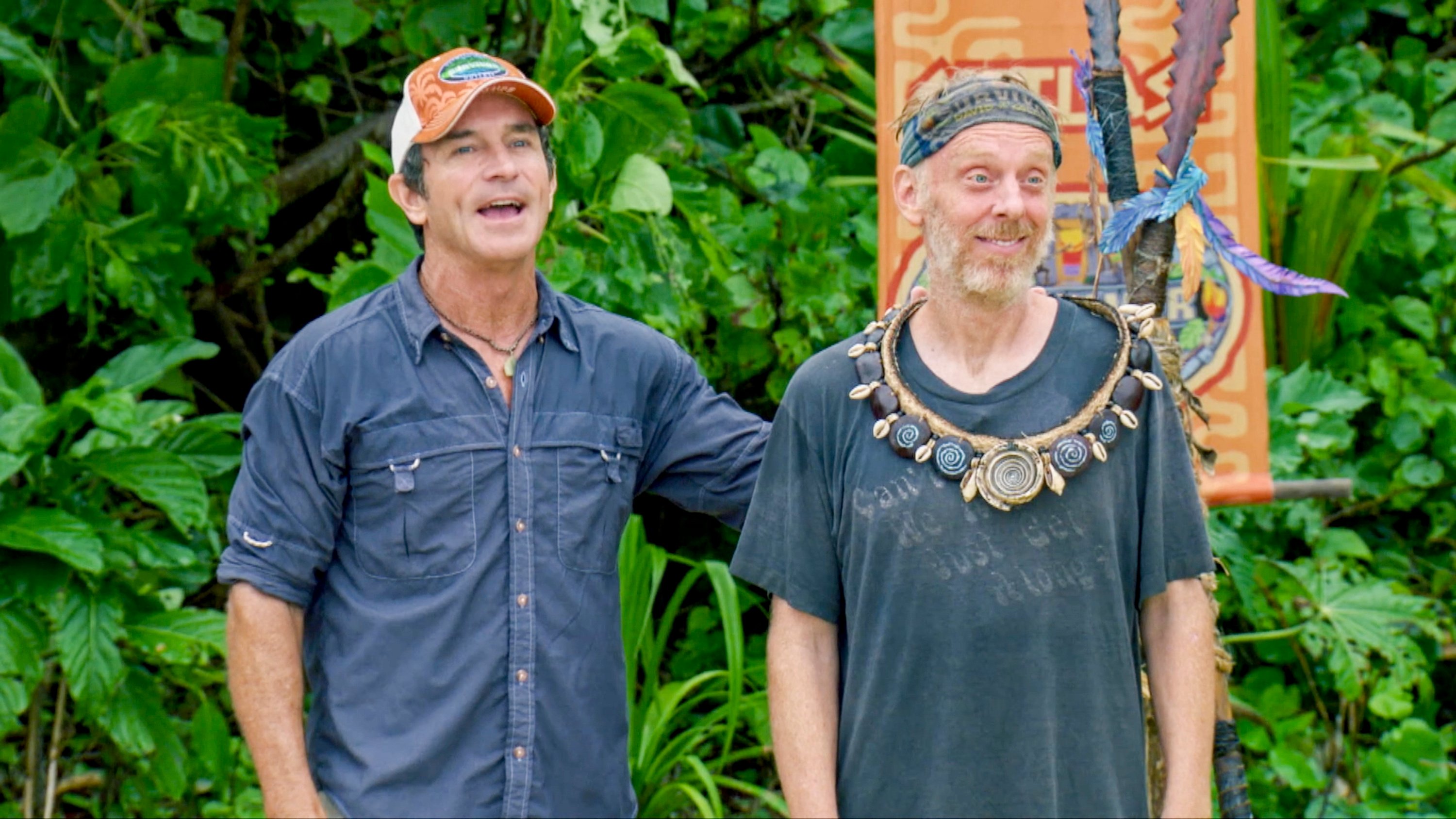 Jeff Probst awards Mike White individual immunity in 'Survivor' Season 37, 'David vs. Goliath.' Jeff wears a dark blue button-up shirt and an orange 'Survivor' hat. Mike wears a faded gray shirt and 'Survivor' buff around his head.