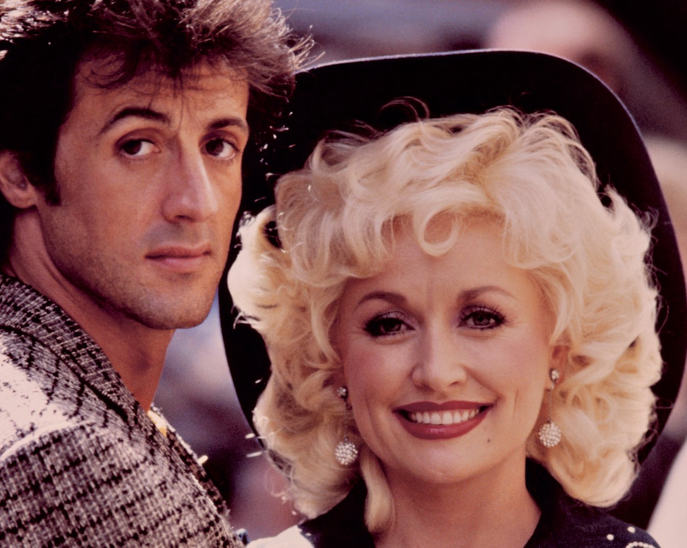 Sylvester Stallone looks at the camera standing next to a smiling Dolly Parton on the set of 'Rhinestone'