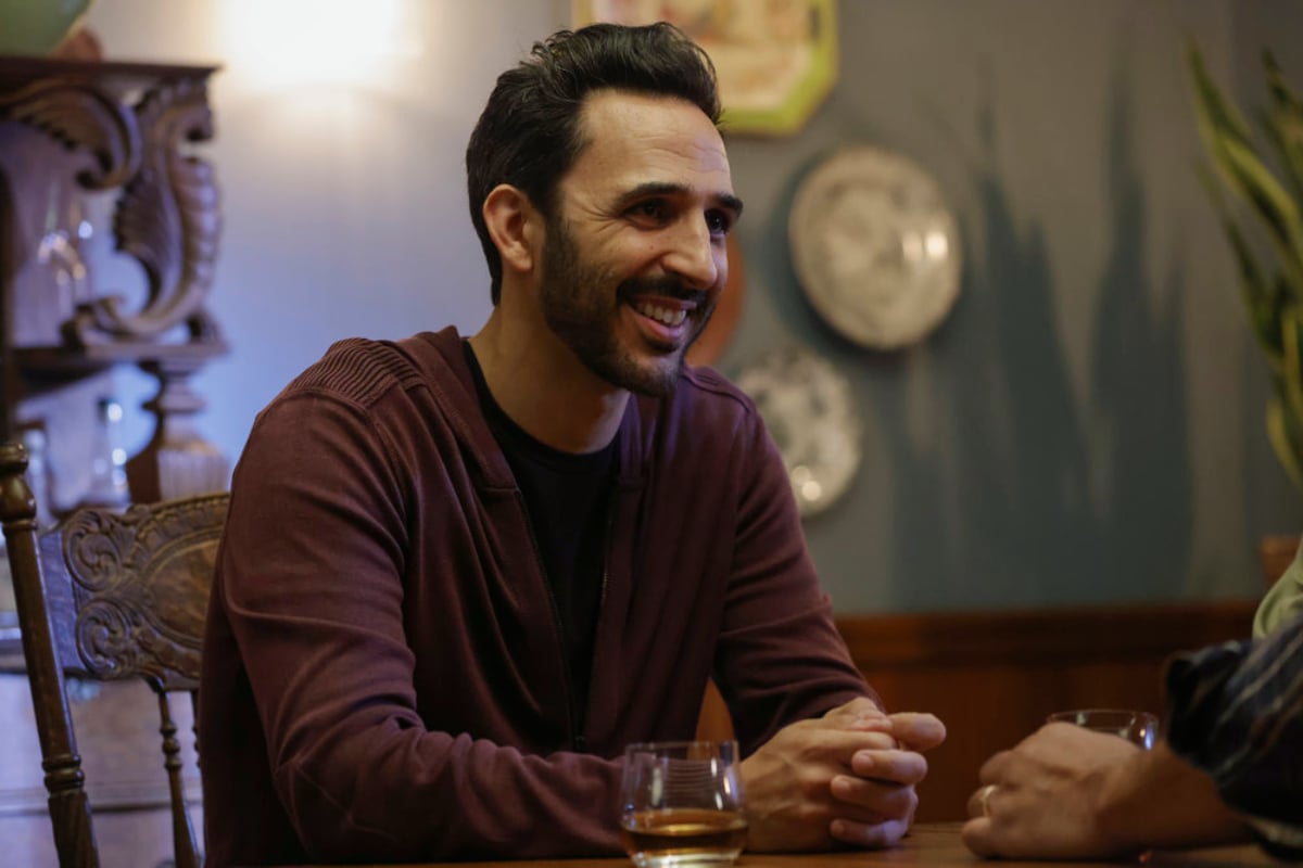 Amir Arison as Aram Mojtabai in The Blacklist Season 9. Aram is smiling and wearing a long-sleeved maroon shirt.