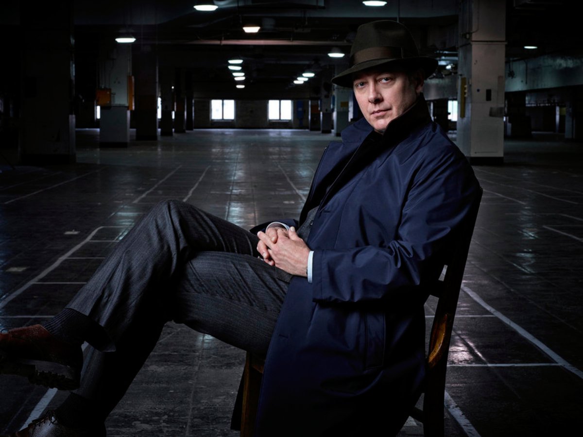 James Spader as Raymon Reddington in The Blacklist is wearing a dark blue coat and black hat and sitting in a chair.