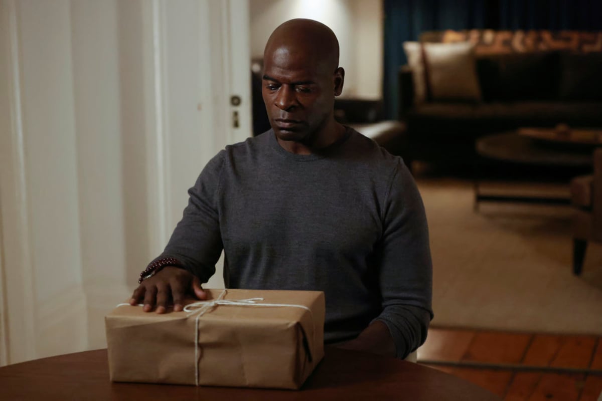 Hisham Tawfiq as Dembe Zuma in The Blacklist'Season 9. Dembe sits at a table with his hand on a wrapped package.
